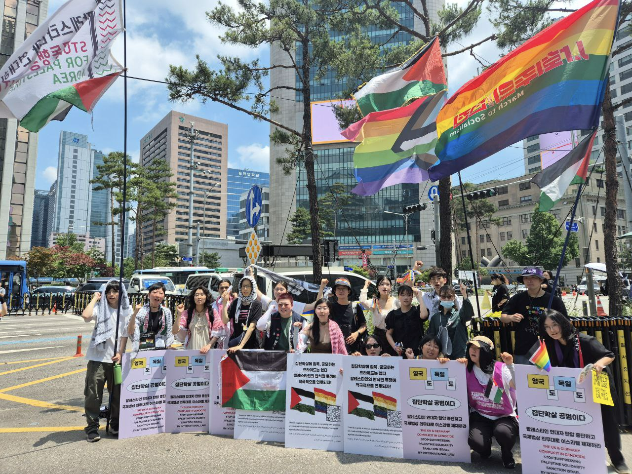 그림 1 QK48이 2025. 6. 14. 서울퀴어문화축제 캠페인을 진행하는 모습. 팔레스타인 국기, 프로그레스 프라이드(성소수자 자긍심) 깃발, ‘영국도 독일도 집단학살 공범이다’ 등의 문구가 적힌 피켓을 든 여러 명의 사람들이 카메라를 향해 투쟁의 의미로 주먹을 쥐고 위로 들어올 리고 있다. (출처 : 성적권리와 재생산정의를 위한 센터 셰어 홈페이지, ‘[활동보고] 퀴어 팔 레스타인 연대의 달 후기’, 2025. 7. 3., https://srhr.kr/announcements/?idx=166477537&bmode=view)