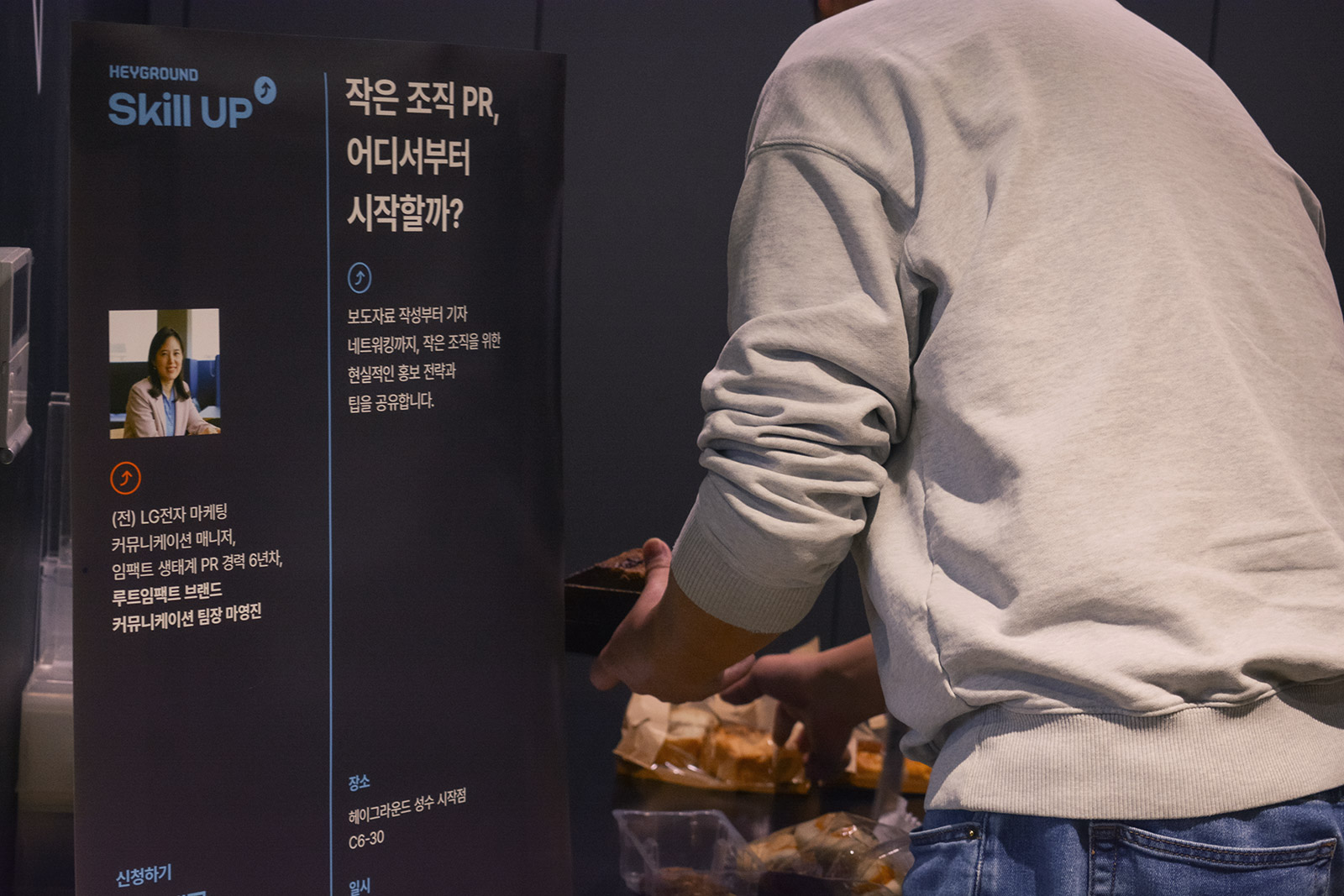 "작은 조직 PR, 어디서부터 시작할까?"라는 주제의 스탠딩 배너 앞에서 한 참가자가 다과를 담고 있다. 배너에는 강연자 마영진 팀장의 이력과 강연 내용이 소개되어 있다.