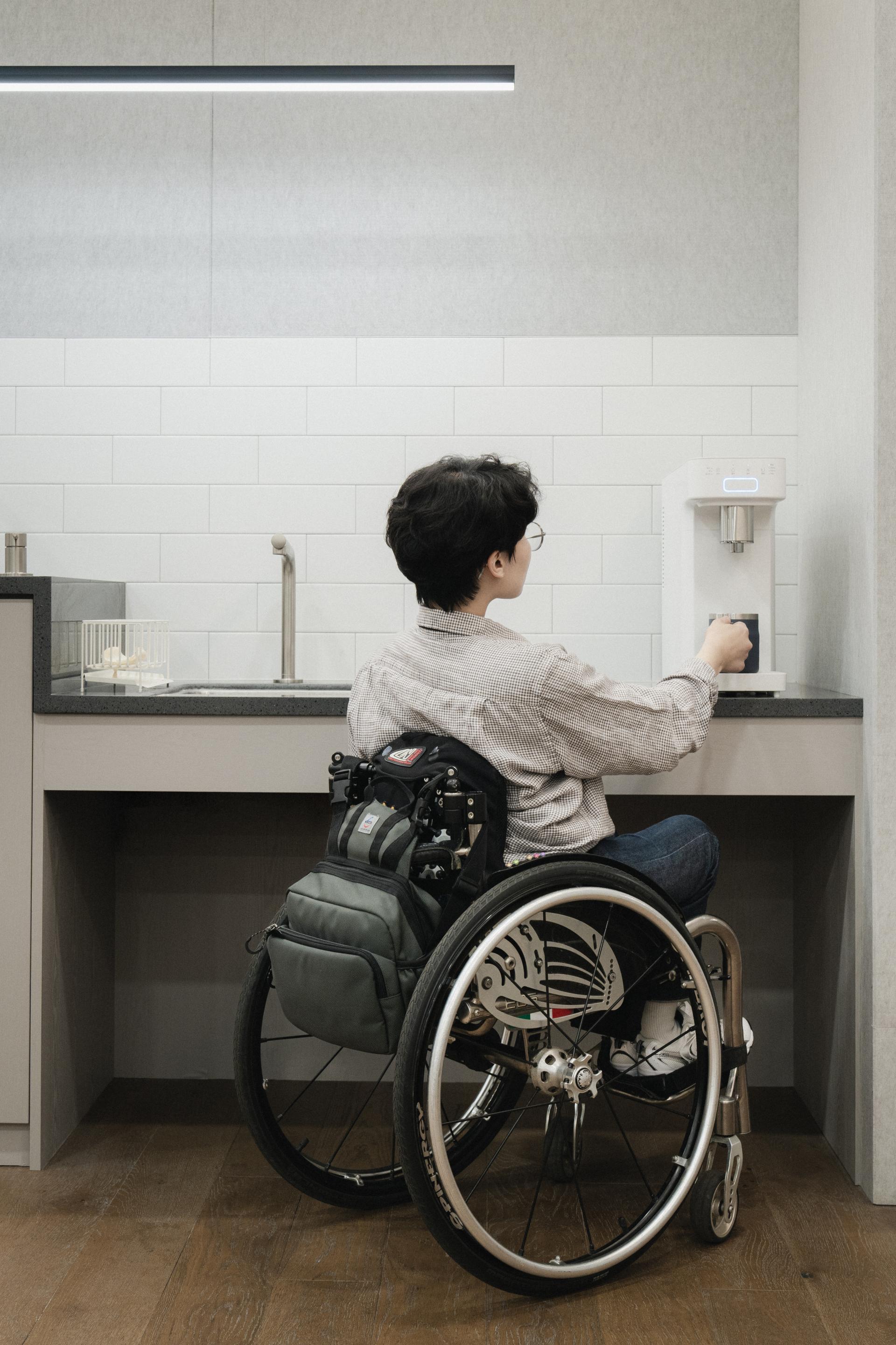 휠체어 사용자가 다양한 높이에 맞춰 설계된 싱크대와 정면 버튼의 정수기를 편리하게 사용하는 모습. 접근성 중심의 공간 디자인을 보여줌.