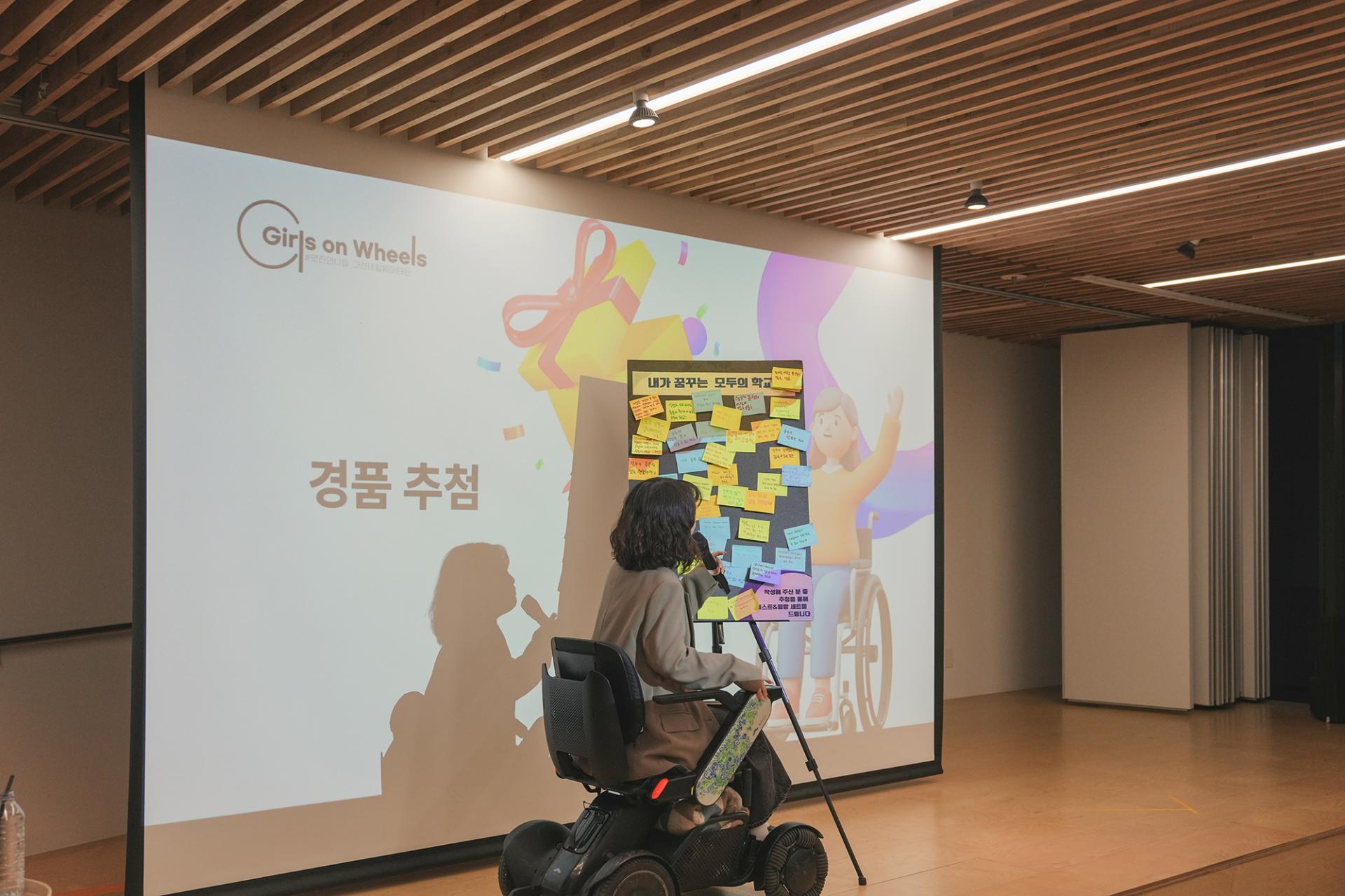 스크린에 경품 추첨이라는 글씨가 써있고 휠체어에 앉은 사회자가 참가자들이 적어 붙인 포스트잇들을 바라보고 있는 모습