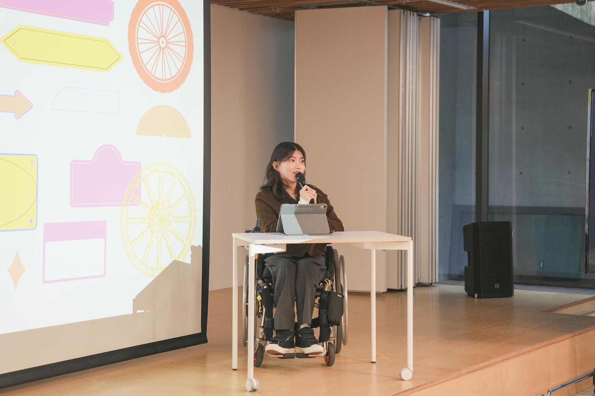 무대에서 발제중인 유지혜님. 짙은 갈색 자켓과 진회색 바지를 입고 휠체어 위에 앉아있다. 발제자 앞에는 흰색 책상이 있고 아이패드가 올려져 있으며 손에는 마이크를 들고있다.