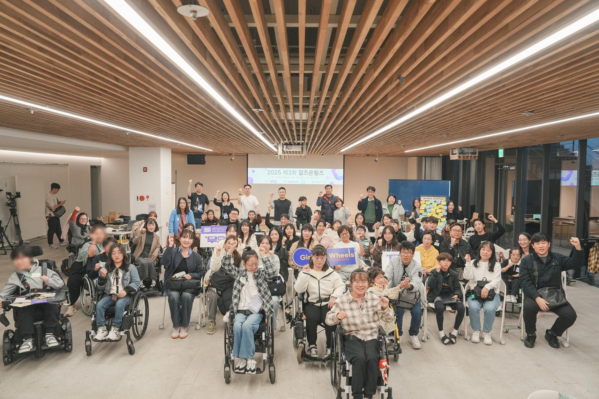 2025 제3회 걸즈온휠즈 행사명이 적힌 스크린을 배경으로 수십 명의 휠체어 이용자들과 비장애인들이 카메라를 바라보며 한 손을 들고 파이팅 포즈를 취하고 있는 단체사진