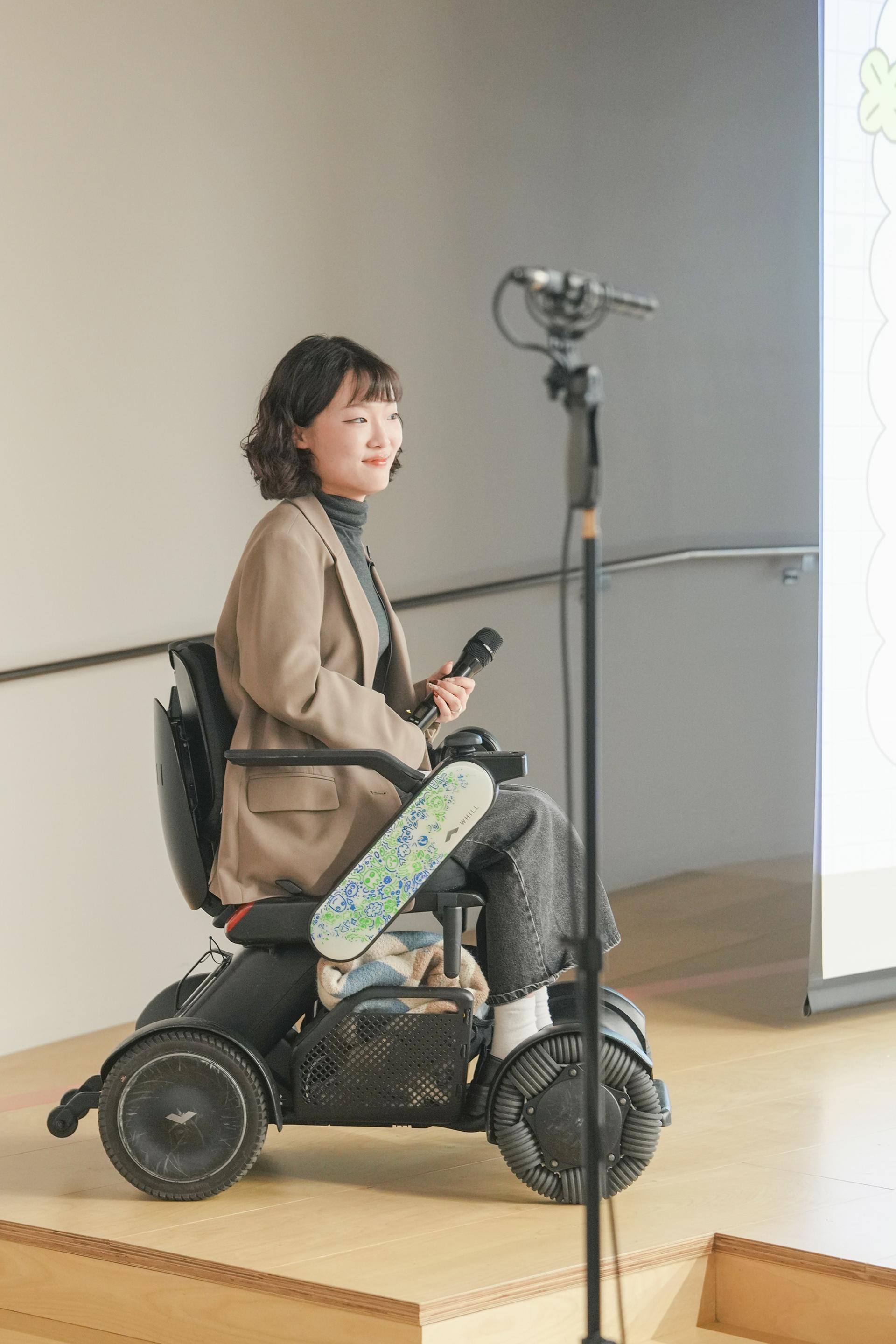 엷은 미소를 띄고 무대에 오른 사회자 김지우님. 갈색 계열 자켓과 짙은 회색의 긴 스커트를 입고 검정색 휠체어에 앉아 왼손에는 마이크를 들고 있다.