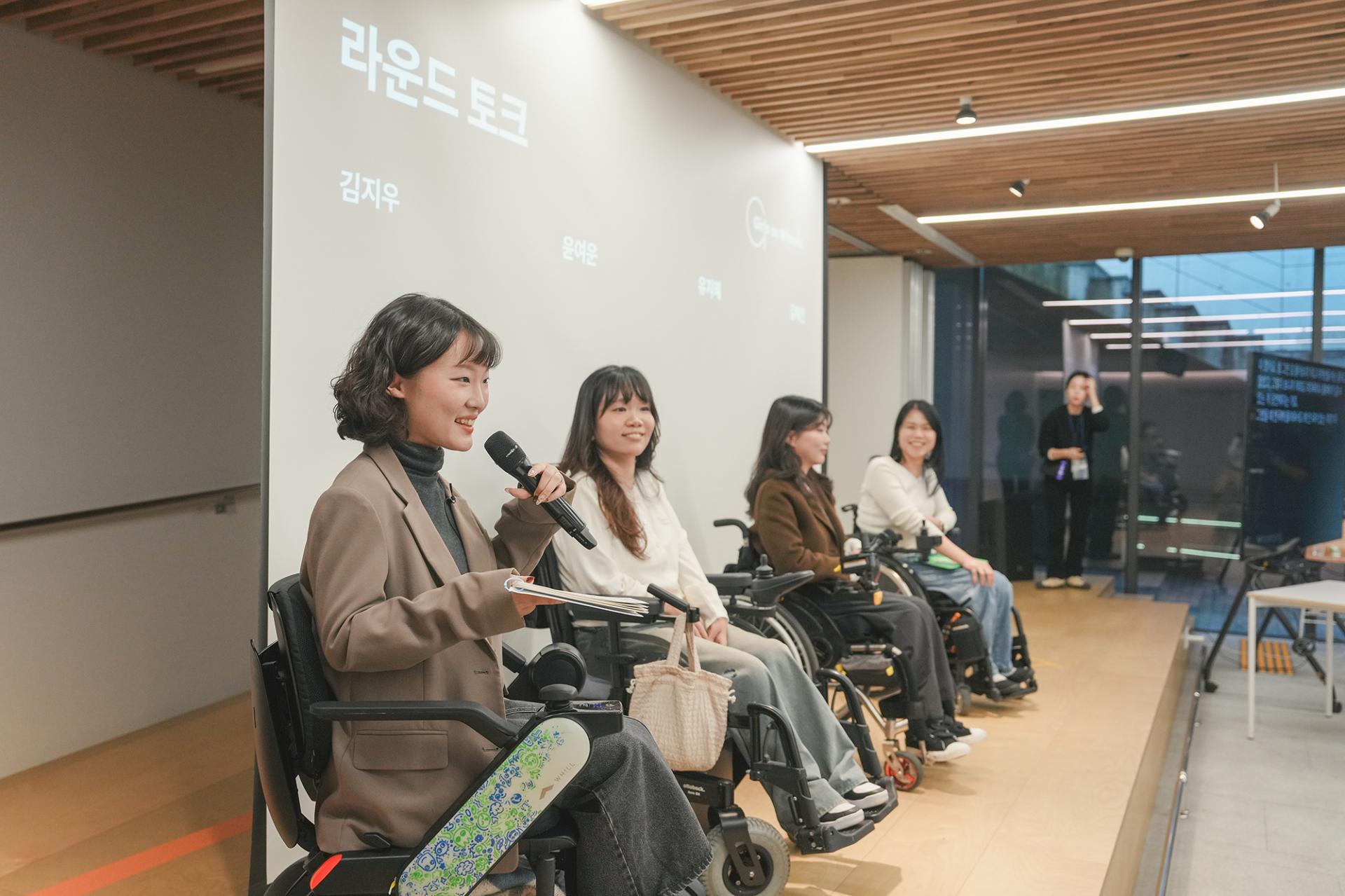 사회자와 세 명의 연사가 무대 위에 나란히 앉아있는 모습. 모두 휠체어에 앉아있고 밝게 웃고있다.