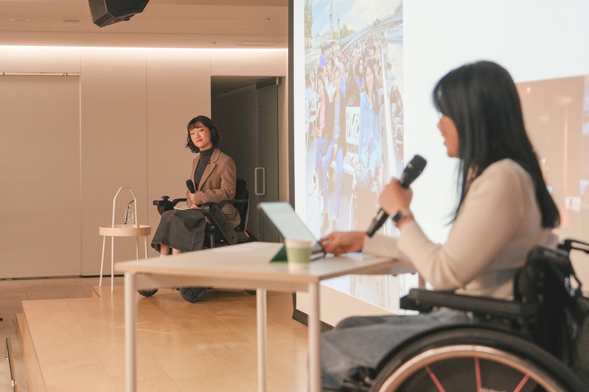 무대 위에서 흰색 옷을 입은 연사가 발표하는 모습을 갈색 상의와 진회색 긴 스커트를 입은 사회자가 바라보고 있다. 사회자와 연사 모두 휠체어에 앉아있다.