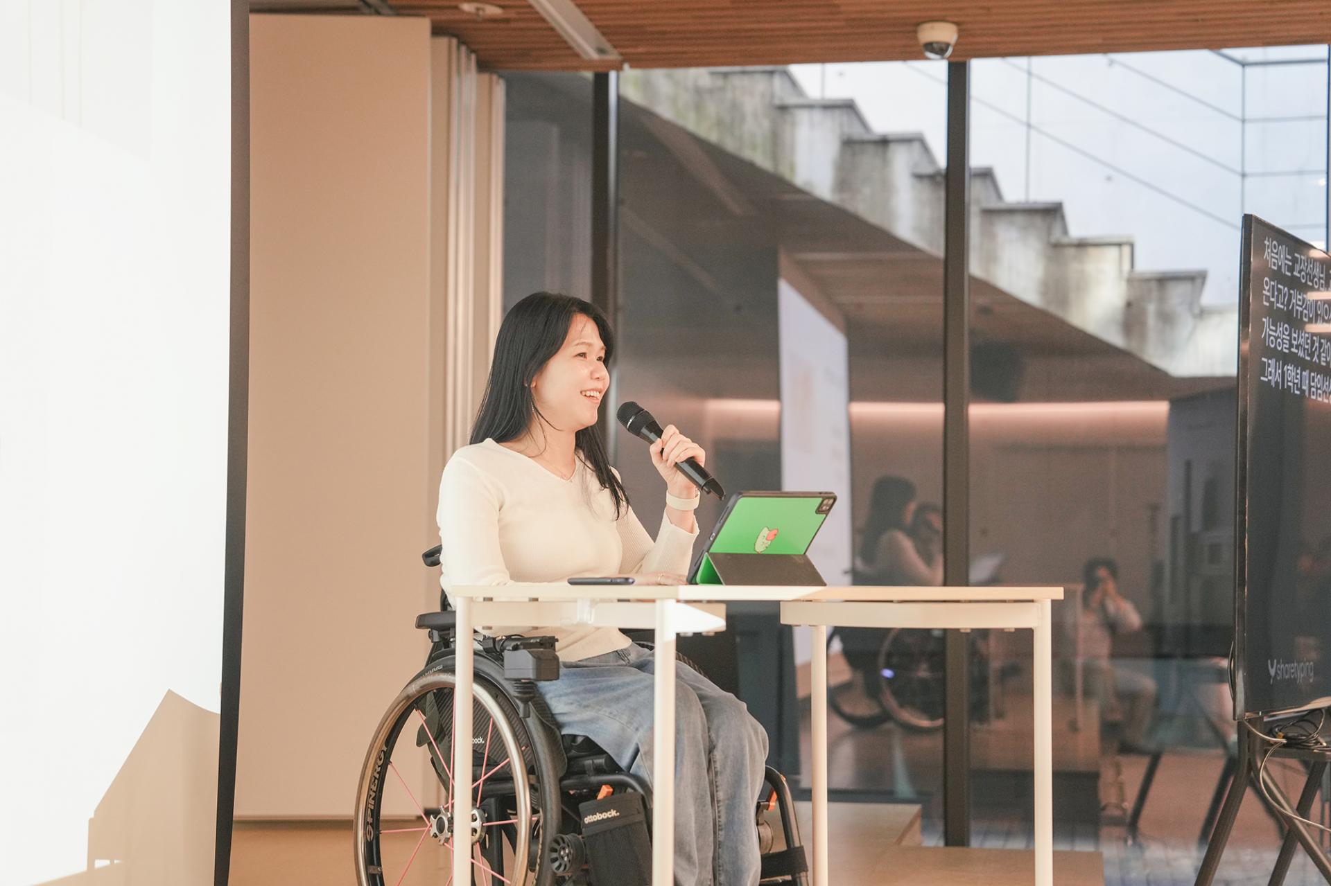 무대에서 발제중인 김혜진님. 흰색 상의와 청바지를 입고 휠체어에 앉아있으며 발제자 앞에는 흰색 책상과 아이패드가 있다. 왼손으로 마이크를 들고 웃으며 말하고 있다.