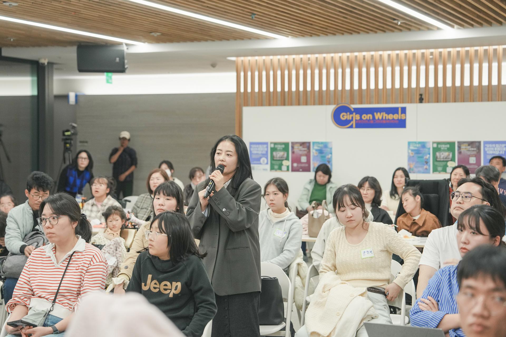 참석자들 사이에서 짙은 회색 자켓을 입은 여성이 일어나 마이크를 들고 질문을 하고있는 모습.