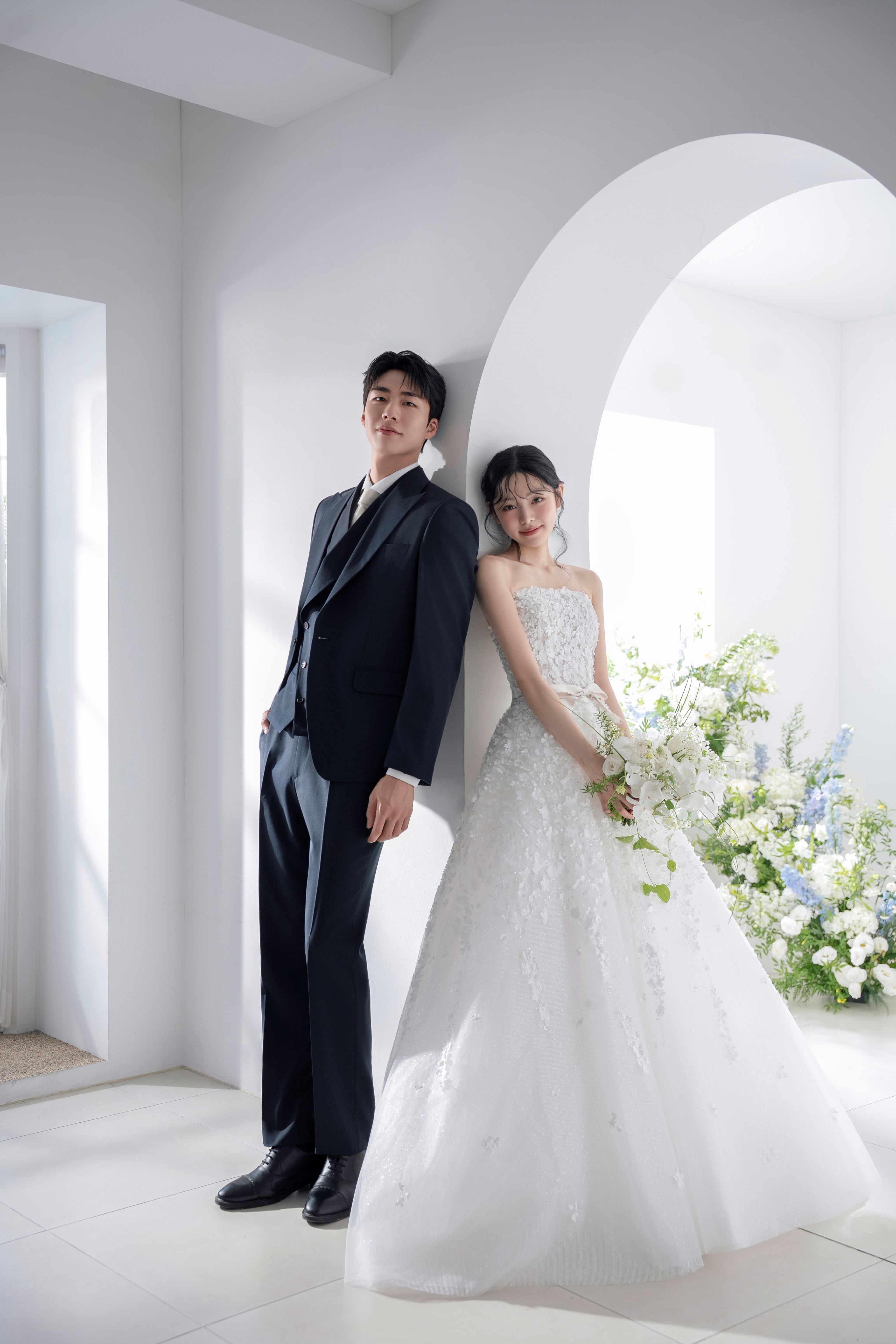 로렌하우스 웨딩 스튜디오 대표 컷 화이트 아치와 화사한 플라워 장식을 배경으로 블랙 수트의 신랑과 입체 플라워 장식의 벨라인 웨딩드레스를 입은 신부가 나란히 기대어 선 모습을 자연광으로 담아낸 밝고 로맨틱한 커플 웨딩 촬영