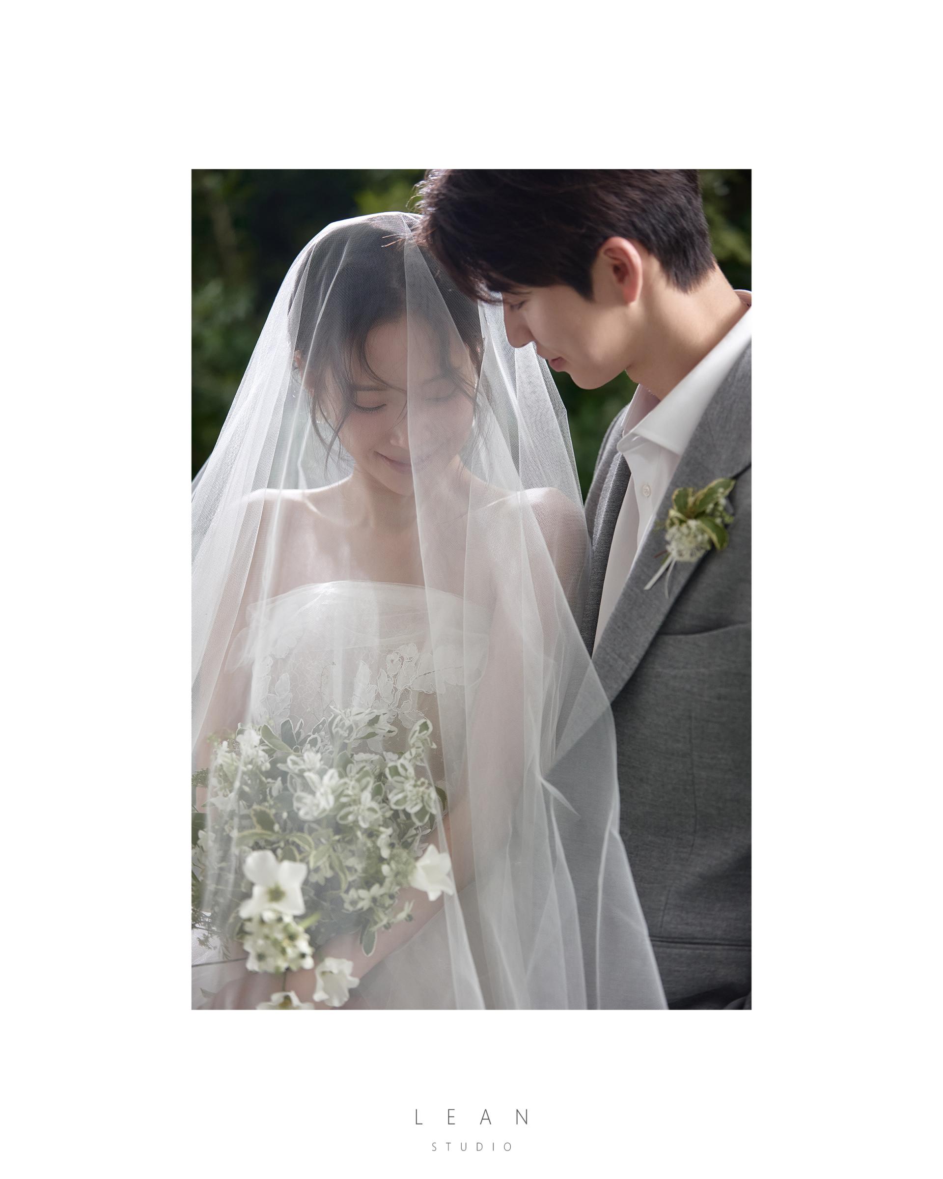 르안스튜디오 웨딩 스튜디오 초록 자연을 배경으로 롱 베일과 화이트 플라워 부케를 든 신부와 그레이 수트 신랑이 가까이 마주 선 모습을 담은 야외 웨딩 촬영컷