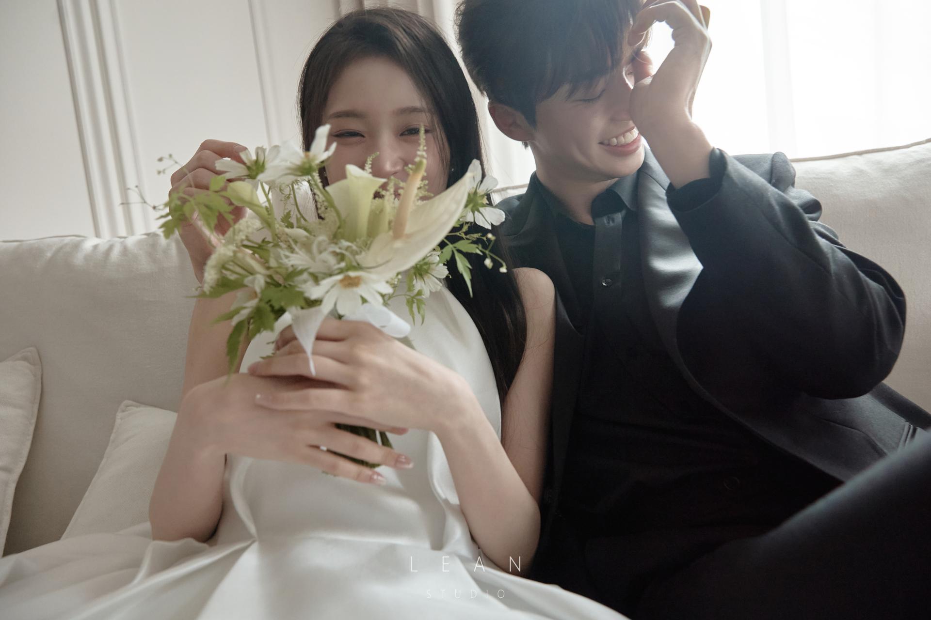 르안스튜디오 웨딩 스튜디오 화이트 소파에 나란히 앉아 화이트 플라워 부케를 들고 환하게 웃는 신랑 신부의 자연스러운 웨딩 촬영컷