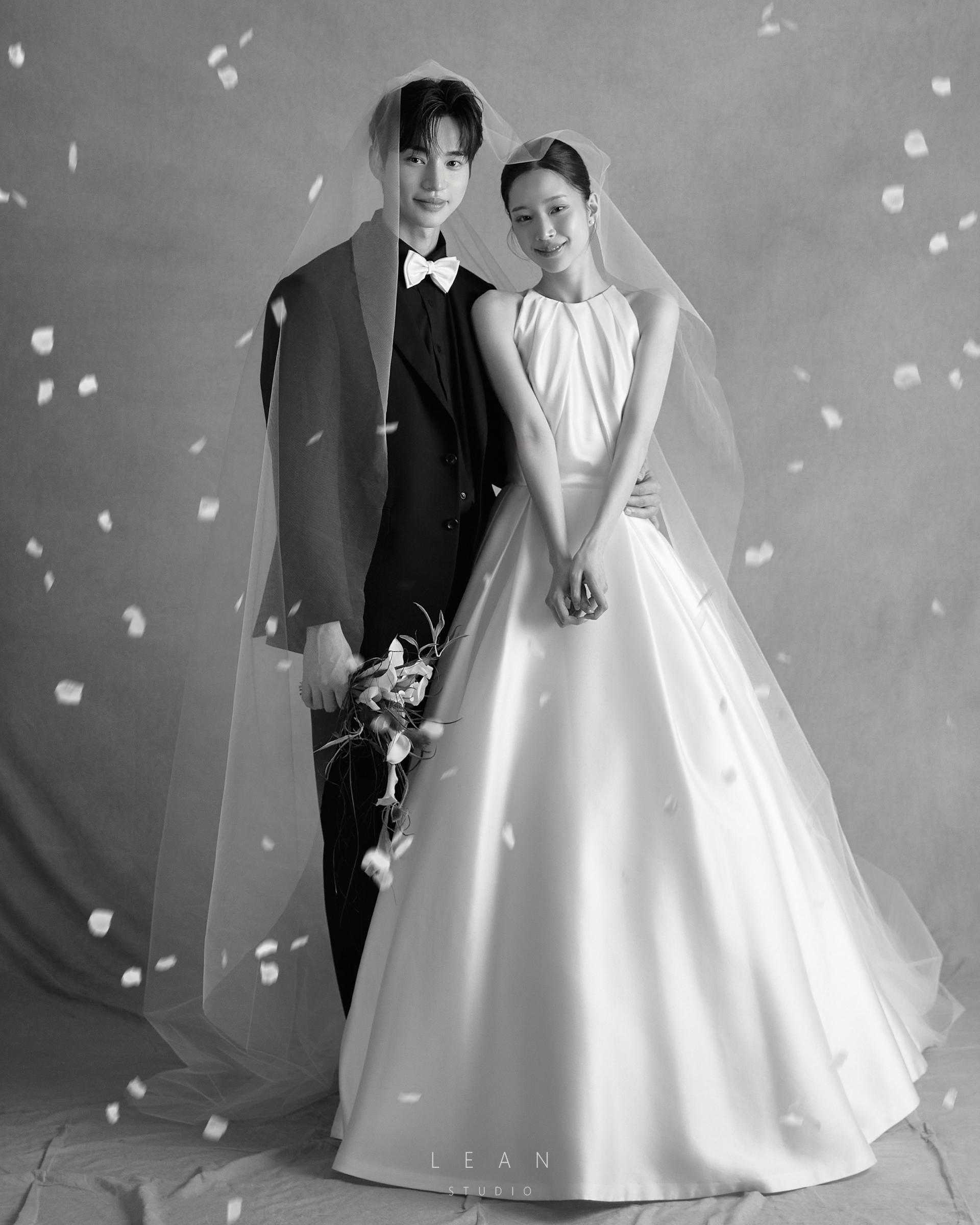 르안 스튜디오 웨딩 스튜디오 롱 베일을 함께 두른 신랑 신부와 흩날리는 꽃잎을 연출하고 클래식한 웨딩드레스와 블랙 턱시도를 담은 흑백 전신 웨딩 촬영컷