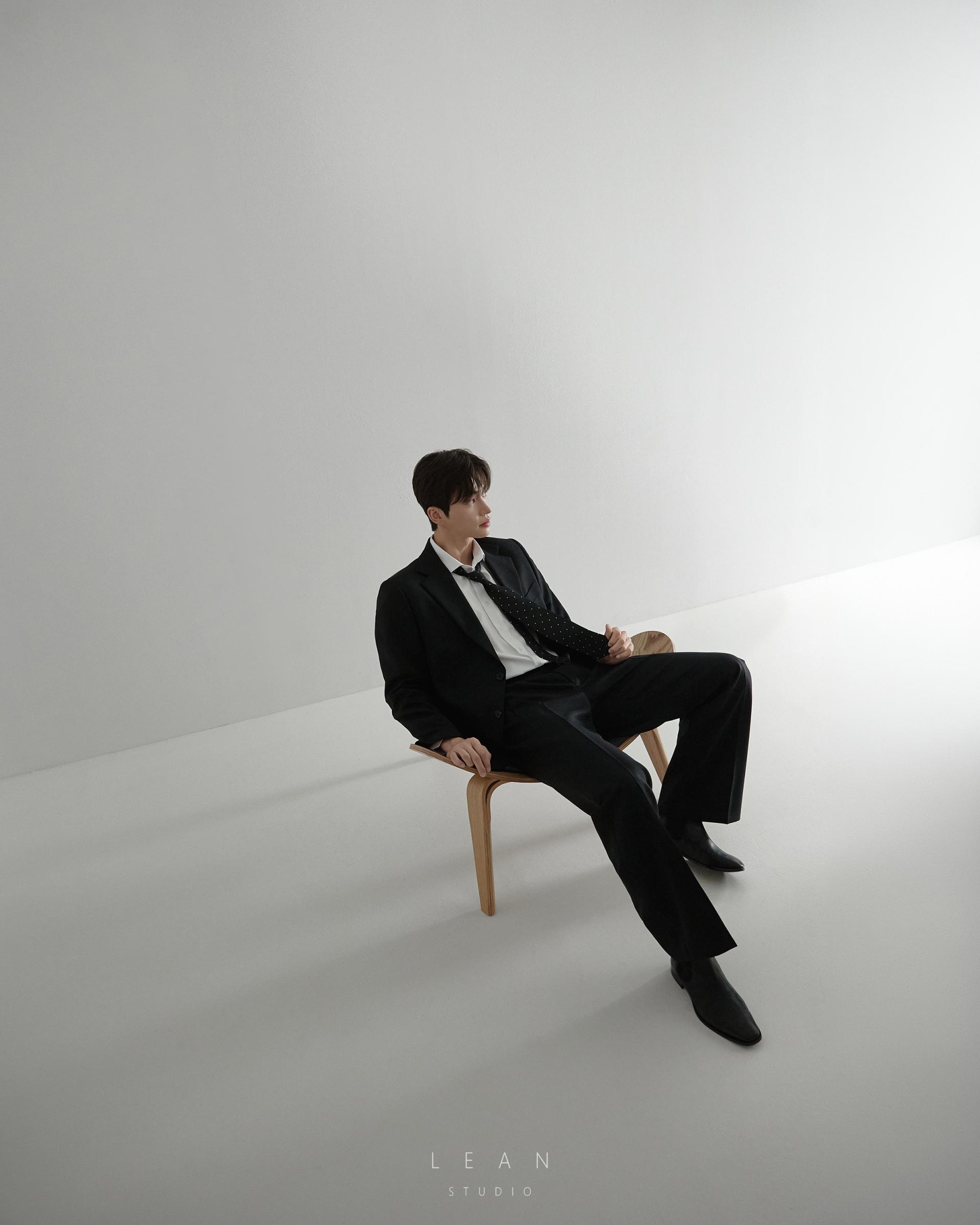 르안스튜디오 웨딩 스튜디오 미니멀한 화이트 공간에서 블랙 수트를 입은 신랑이 우드 체어에 앉아 여백을 살린 구도의 신랑 단독 웨딩 촬영컷