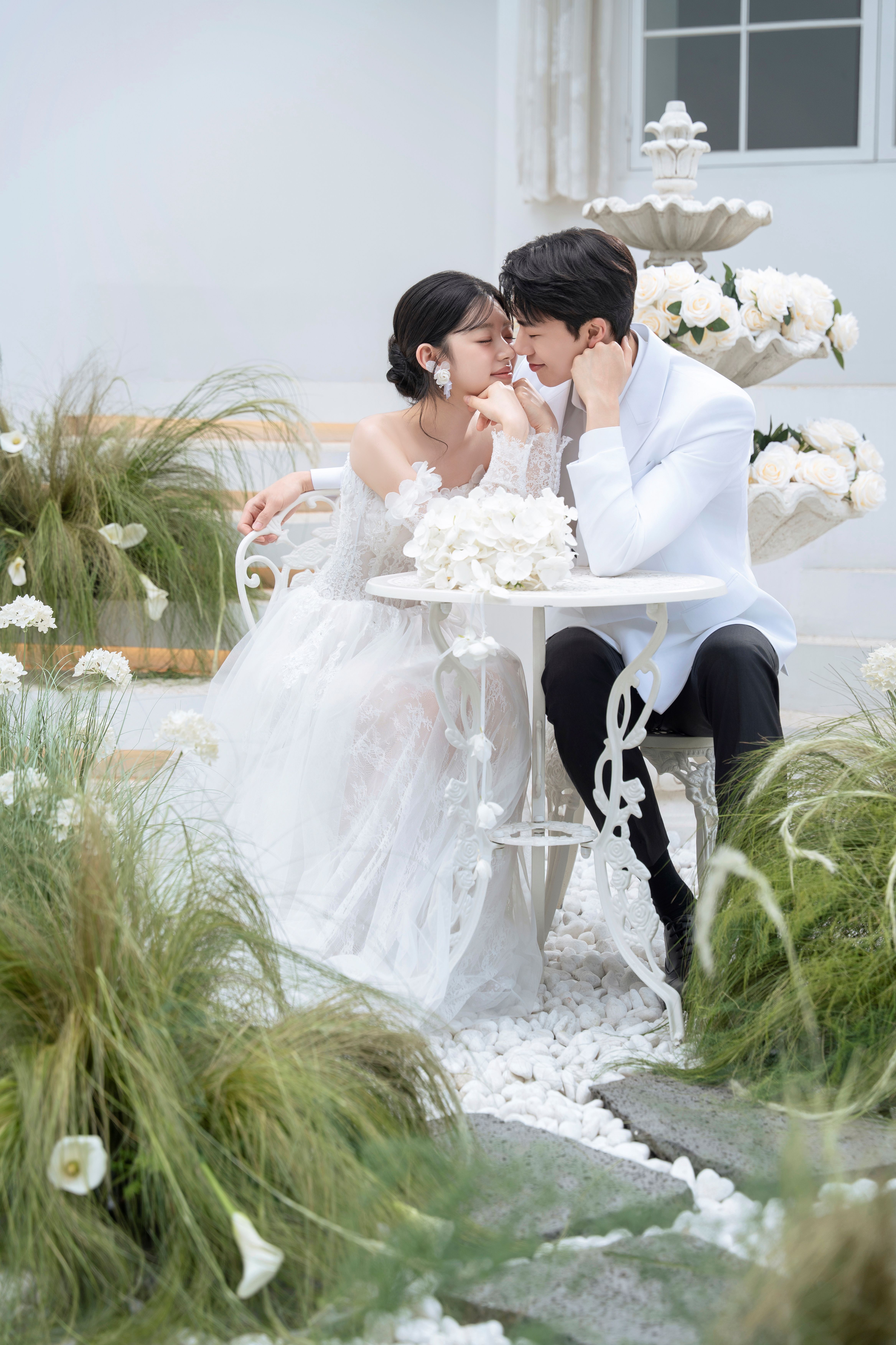 로렌하우스 웨딩 스튜디오 대표 컷 화이트 플라워와 분수 오브제가 어우러진 유럽풍 가든 세트에서 화이트 재킷의 신랑과 레이스 웨딩드레스의 신부가 테이블에 마주 앉아 얼굴을 맞댄 모습을 담아낸 로맨틱하고 화사한 야외 웨딩 촬영