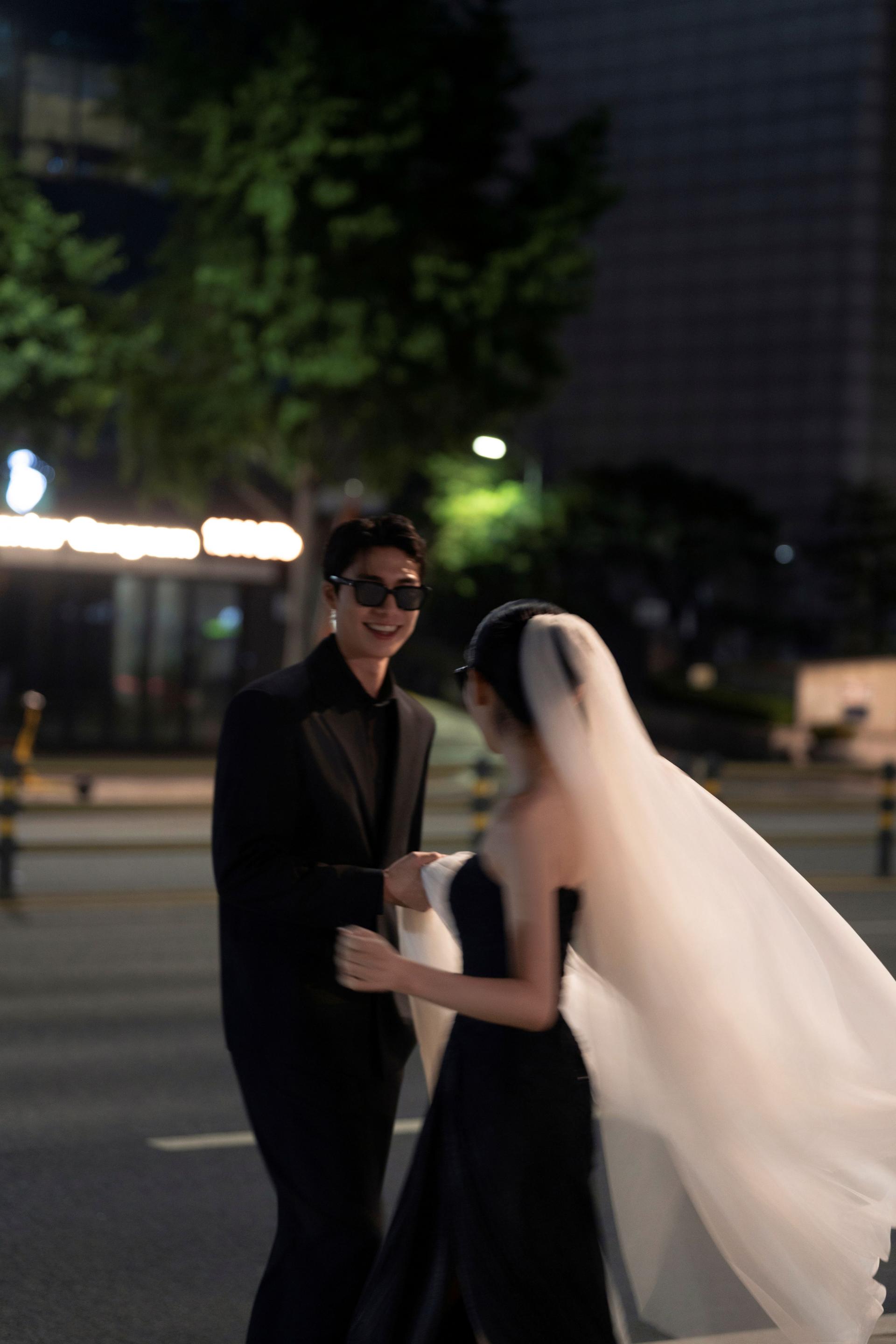 로렌하우스 웨딩 스튜디오 야외 밤거리에서 선글라스를 끼고 블랙 드레스와 수트를 입은 신랑 신부가 마주 보며 미소 짓는 모습을 담은 커플 웨딩 사진