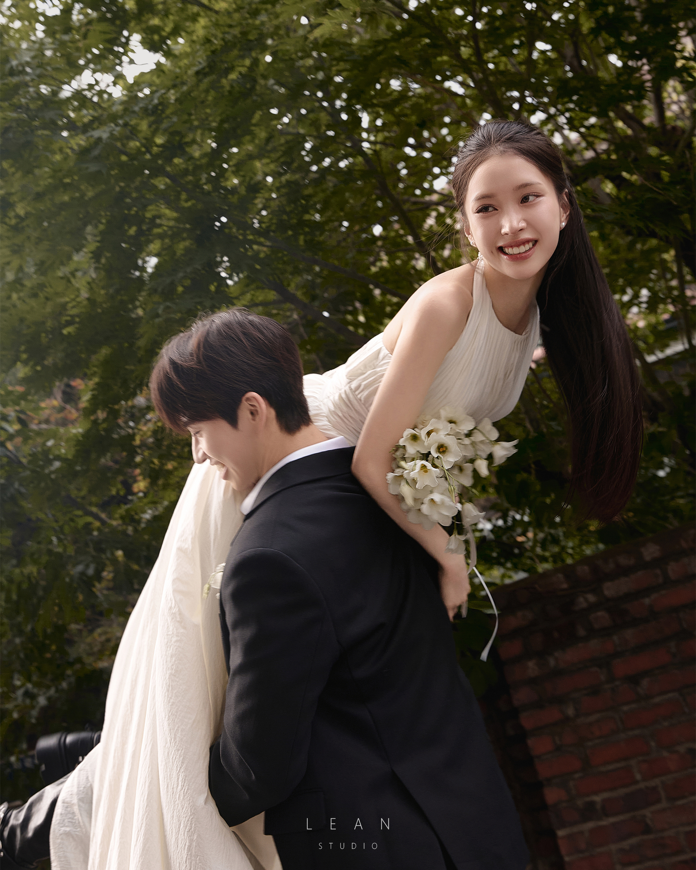 르안 웨딩 스튜디오 화이트 배경에서 블랙 수트 신랑과 레이스 웨딩드레스 신부가 핑크 플라워 부케를 사이에 두고 다정하게 얼굴을 맞댄 플라워 콘셉트 웨딩 촬영컷