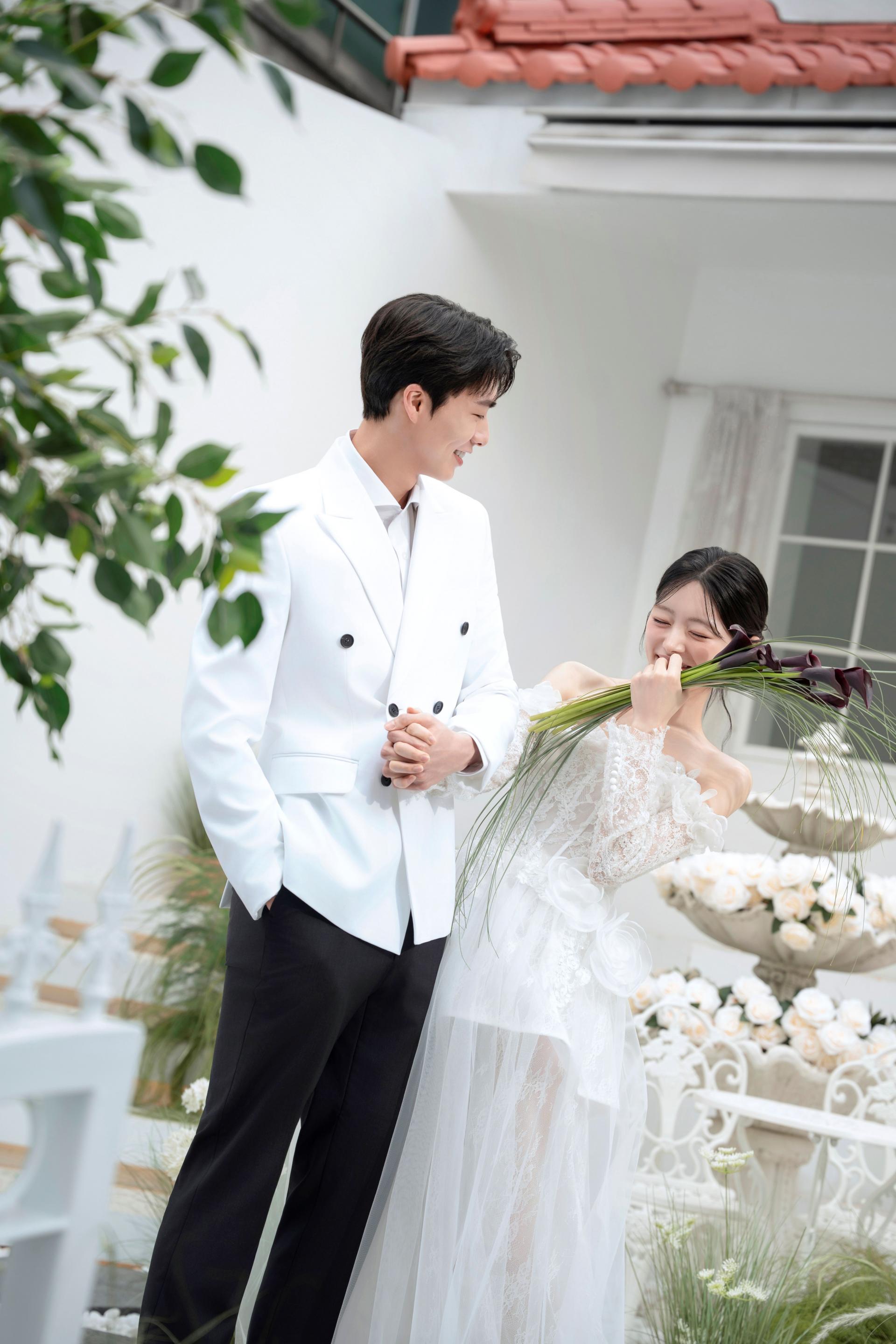 로렌하우스 웨딩 스튜디오 화이트 수트를 입은 신랑과 레이스 드레스를 입고 부케를 든 신부가 야외 정원에서 마주보며 웃고 있는 커플 웨딩 사진