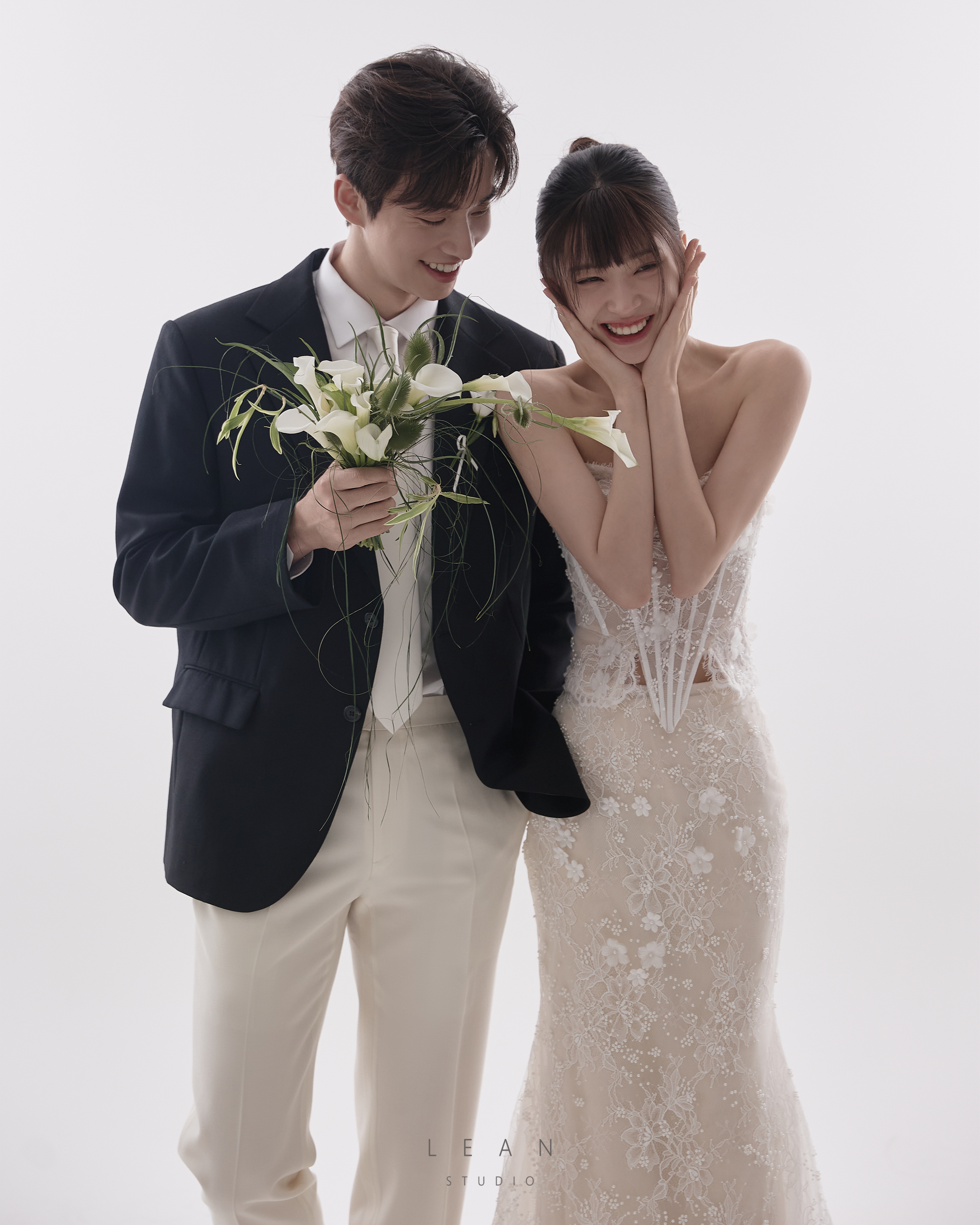 르안 웨딩 스튜디오 화이트 배경에서 블랙 재킷과 화이트 팬츠를 입은 신랑이 화이트 부케를 들고 레이스 슬림 웨딩드레스의 신부와 함께 밝게 웃는 웨딩 촬영컷