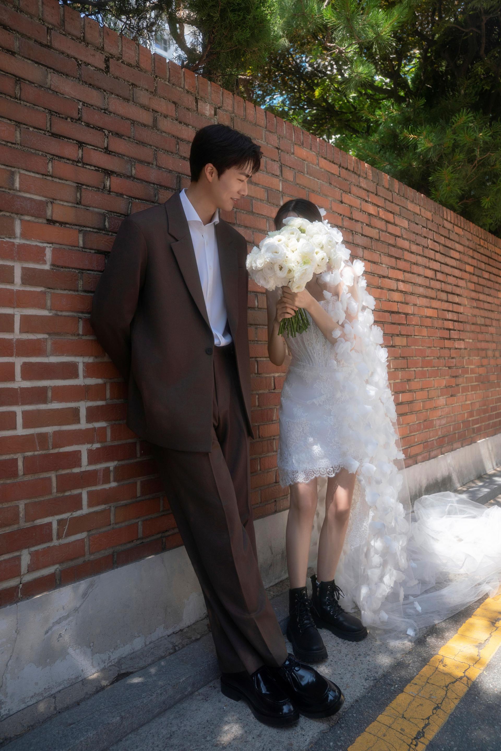 로렌하우스 웨딩 스튜디오 붉은 벽돌 담장을 배경으로 브라운 수트를 입은 신랑과 화이트 부케로 얼굴을 가린 신부가 서 있는 야외 웨딩 사진