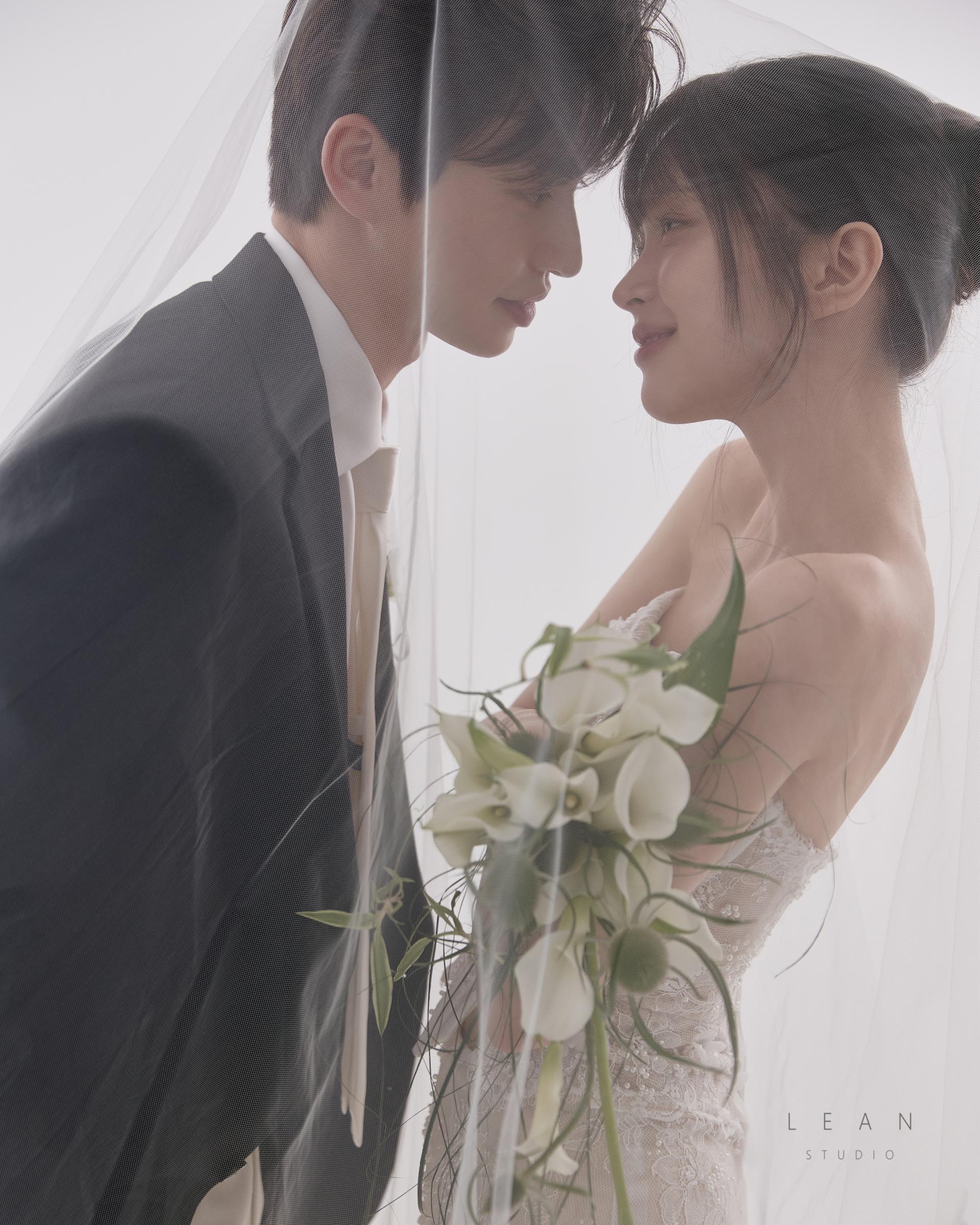 르안스튜디오 웨딩 스튜디오 롱 베일 아래에서 서로를 마주 보는 신랑 신부와 화이트 카라 부케를 함께 담은 로맨틱한 클로즈업 웨딩 촬영컷