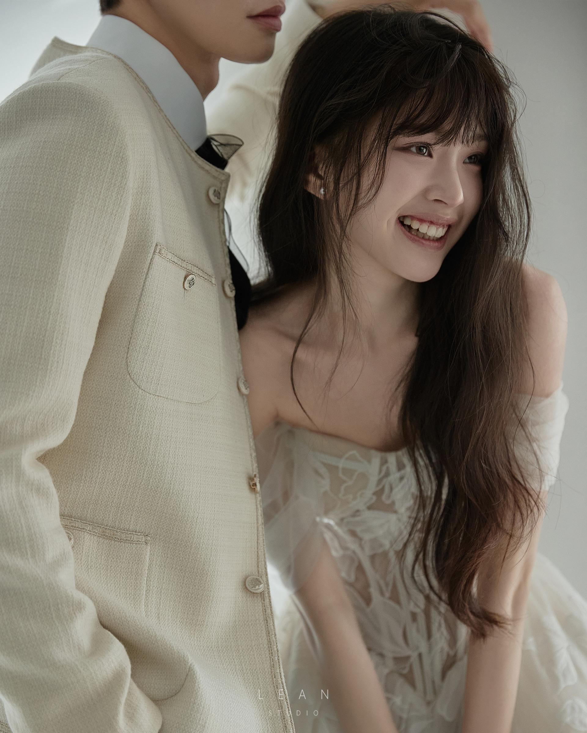 르안 스튜디오 웨딩 스튜디오 곡선형 아치와 창가 자연광이 어우러진 화이트 공간에서 플라워 자수 웨딩드레스와 아이보리 재킷을 입고 나란히 선 신랑 신부 전신 웨딩 촬영컷