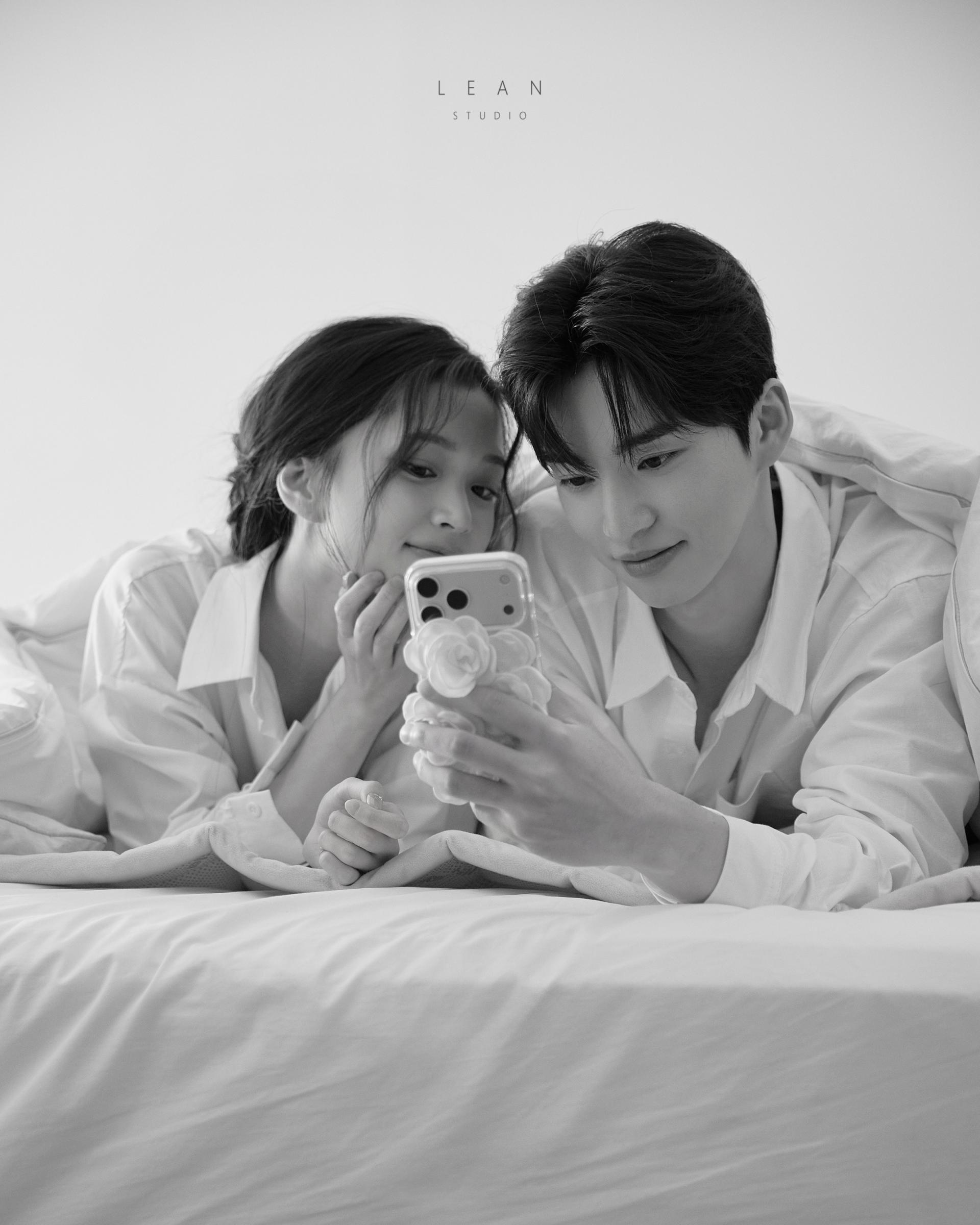 르안 스튜디오 웨딩 스튜디오 화이트 셔츠를 입은 신랑 신부가 침대에 나란히 엎드려 스마트폰을 함께 바라보는 자연스러운 일상을 연출한 흑백 커플 웨딩 촬영컷