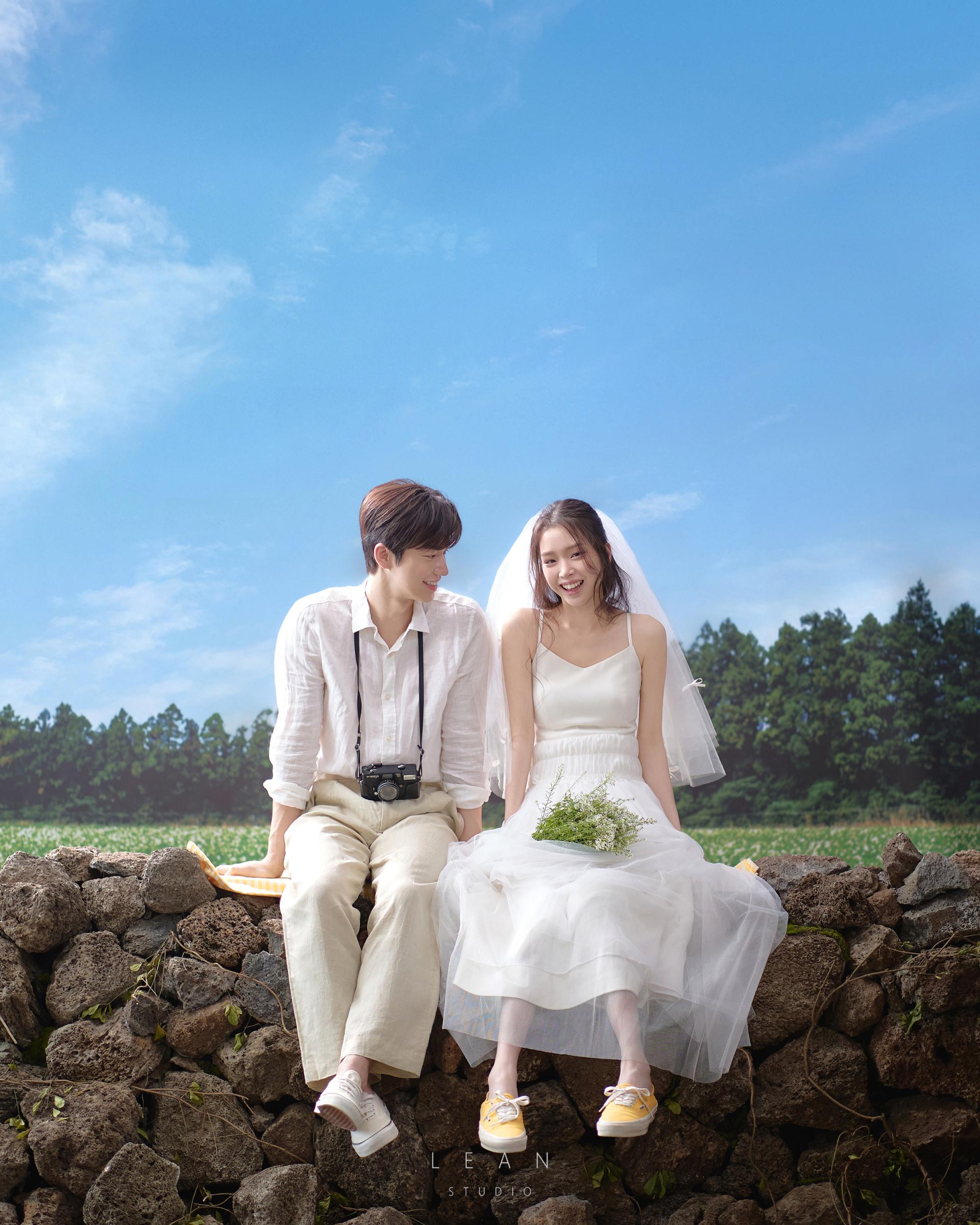 르안스튜디오 웨딩 스튜디오 푸른 하늘과 초록 들판을 배경으로 돌담에 나란히 앉아 카메라를 멘 신랑과 베일을 쓴 신부가 밝게 웃는 자연스러운 야외 웨딩 촬영컷