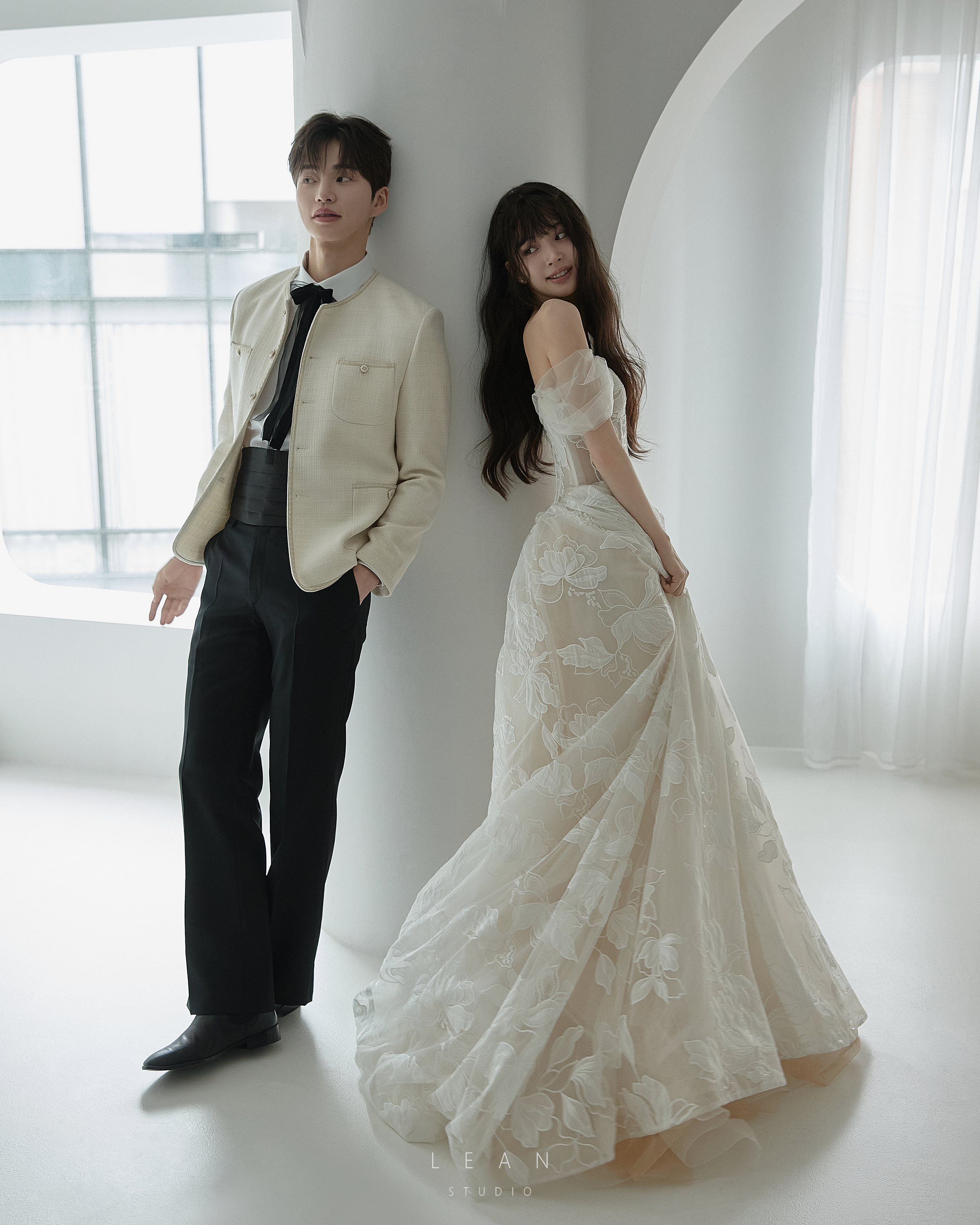 르안 웨딩 스튜디오 자연광이 들어오는 화이트 아치형 공간에서 아이보리 재킷과 블랙 팬츠를 입은 신랑과 입체 플라워 자수 웨딩드레스를 입은 신부가 나란히 포즈를 취한 웨딩 촬영컷