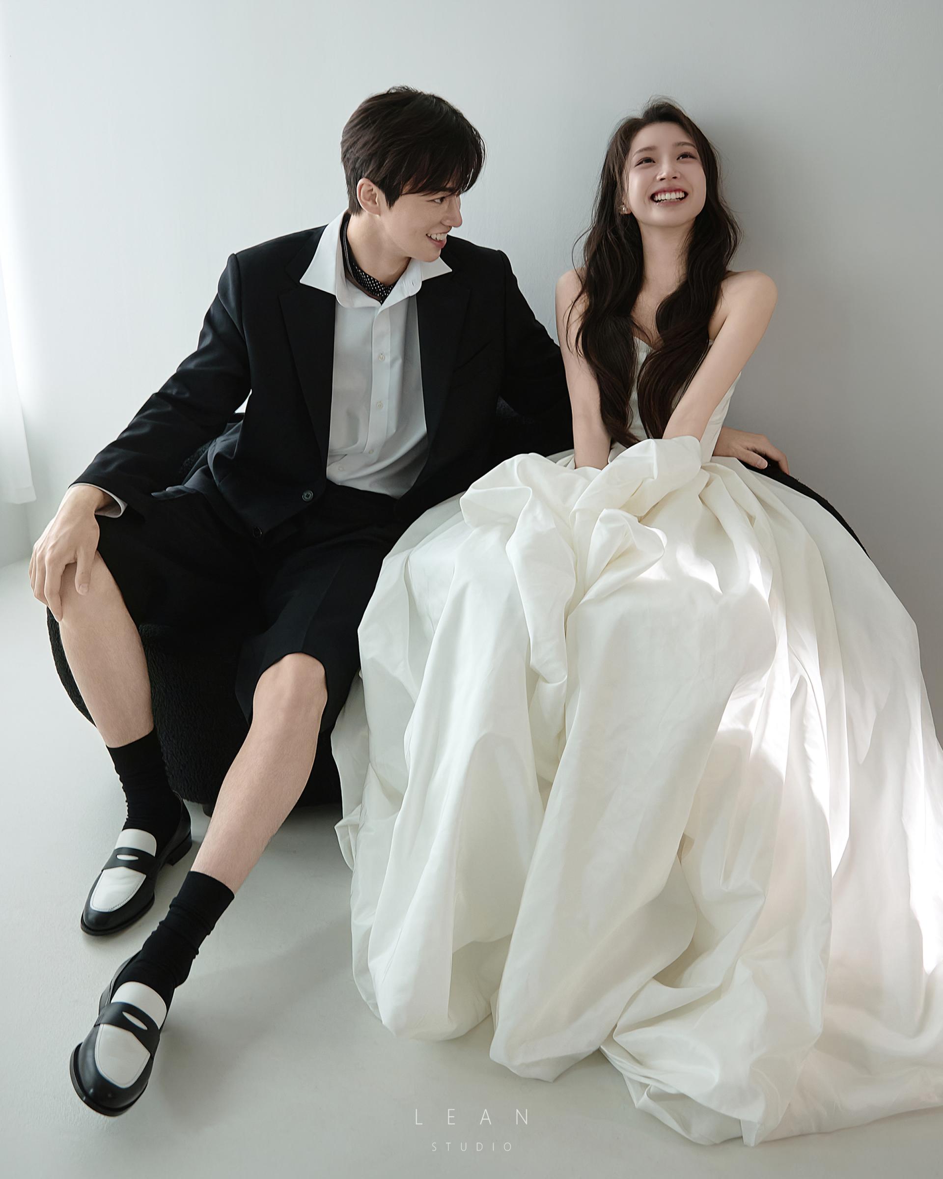 르안 웨딩 스튜디오 화이트 공간에서 블랙 수트와 하프 팬츠를 입은 신랑이 풍성한 화이트 웨딩드레스를 입고 환하게 웃는 신부를 바라보며 나란히 앉아 있는 자연스러운 웨딩 촬영컷