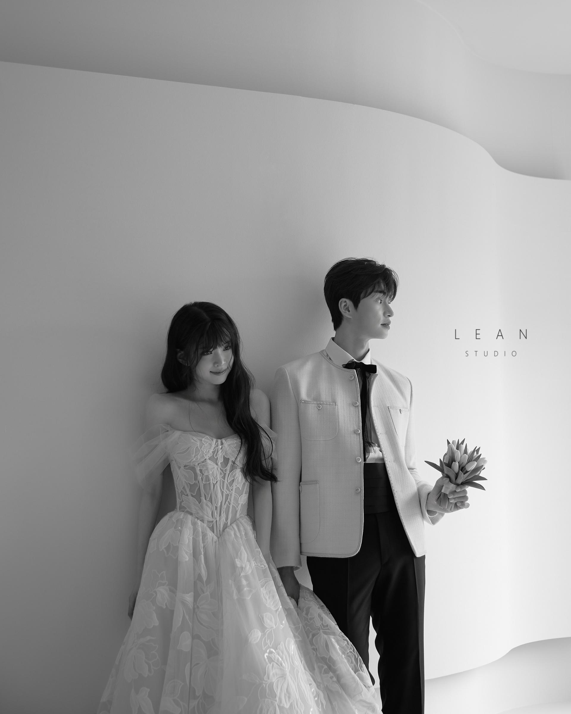 르안스튜디오 웨딩 스튜디오 곡선형 화이트 배경 앞에서 플라워 자수 오프숄더 드레스를 입은 신부와 부케를 든 신랑을 담은 흑백 웨딩 촬영컷