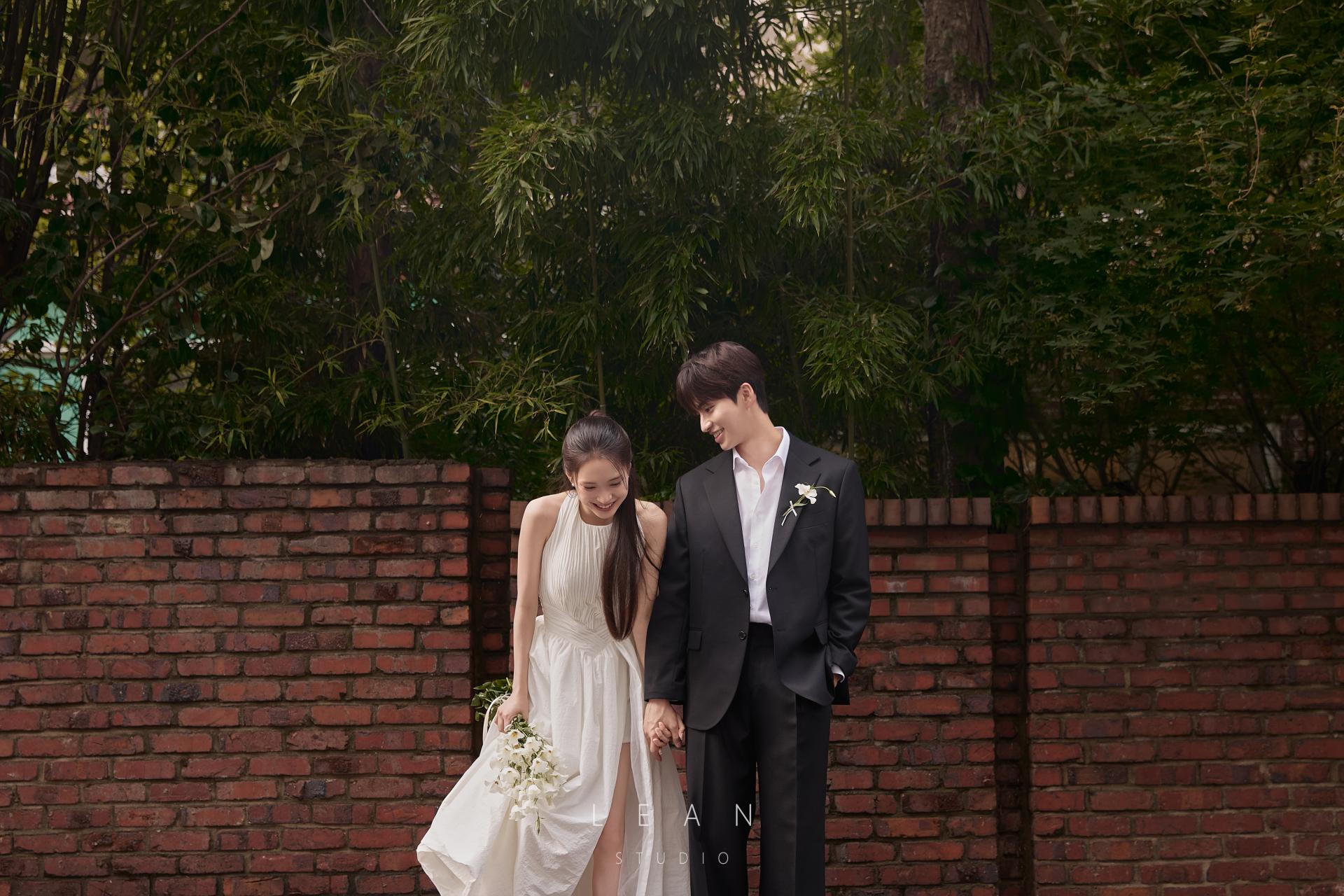 르안스튜디오 웨딩 스튜디오 붉은 벽돌과 울창한 녹음을 배경으로 화이트 드레스와 블랙 수트를 입고 손을 잡으며 웃는 신랑 신부의 야외 웨딩 촬영컷