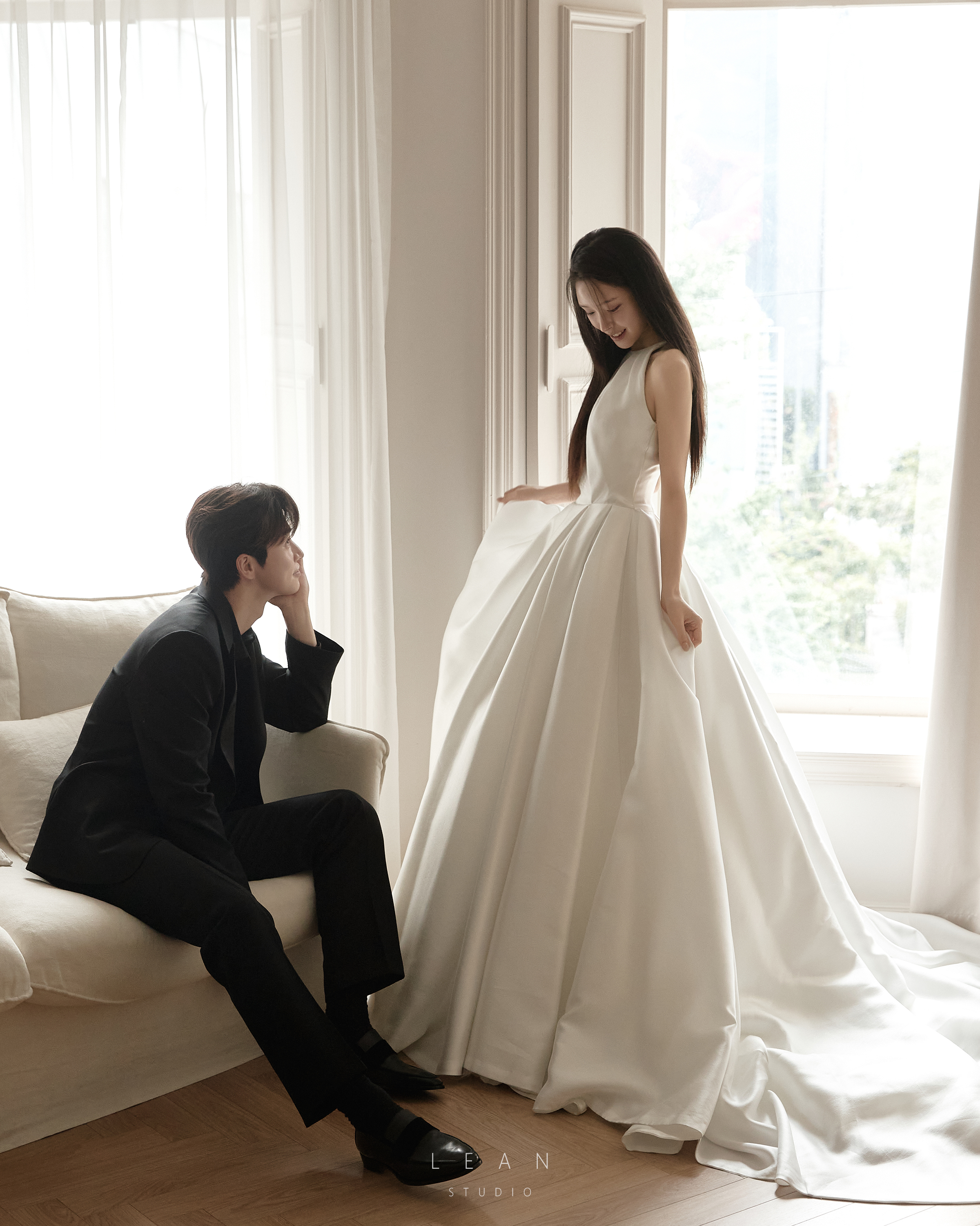 르안 웨딩 스튜디오 자연광이 들어오는 화이트 창가에서 블랙 수트 신랑이 소파에 앉아 신부를 바라보고 민소매 실크 볼륨 웨딩드레스를 입은 신부가 마주 선 웨딩 촬영컷