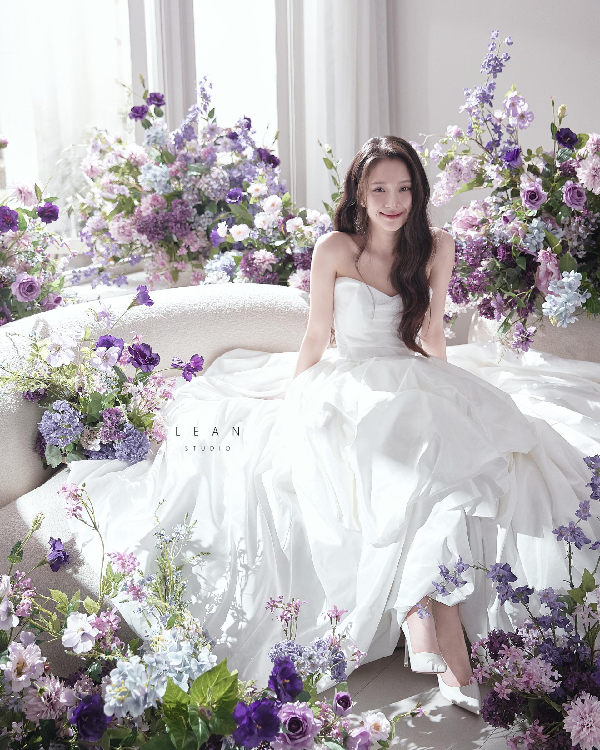 르안스튜디오 웨딩 스튜디오 퍼플과 라벤더 컬러 플라워로 장식된 창가 소파에서 풍성한 화이트 웨딩드레스를 펼치고 앉아 미소 짓는 신부의 자연광 플라워씬 웨딩 촬영컷