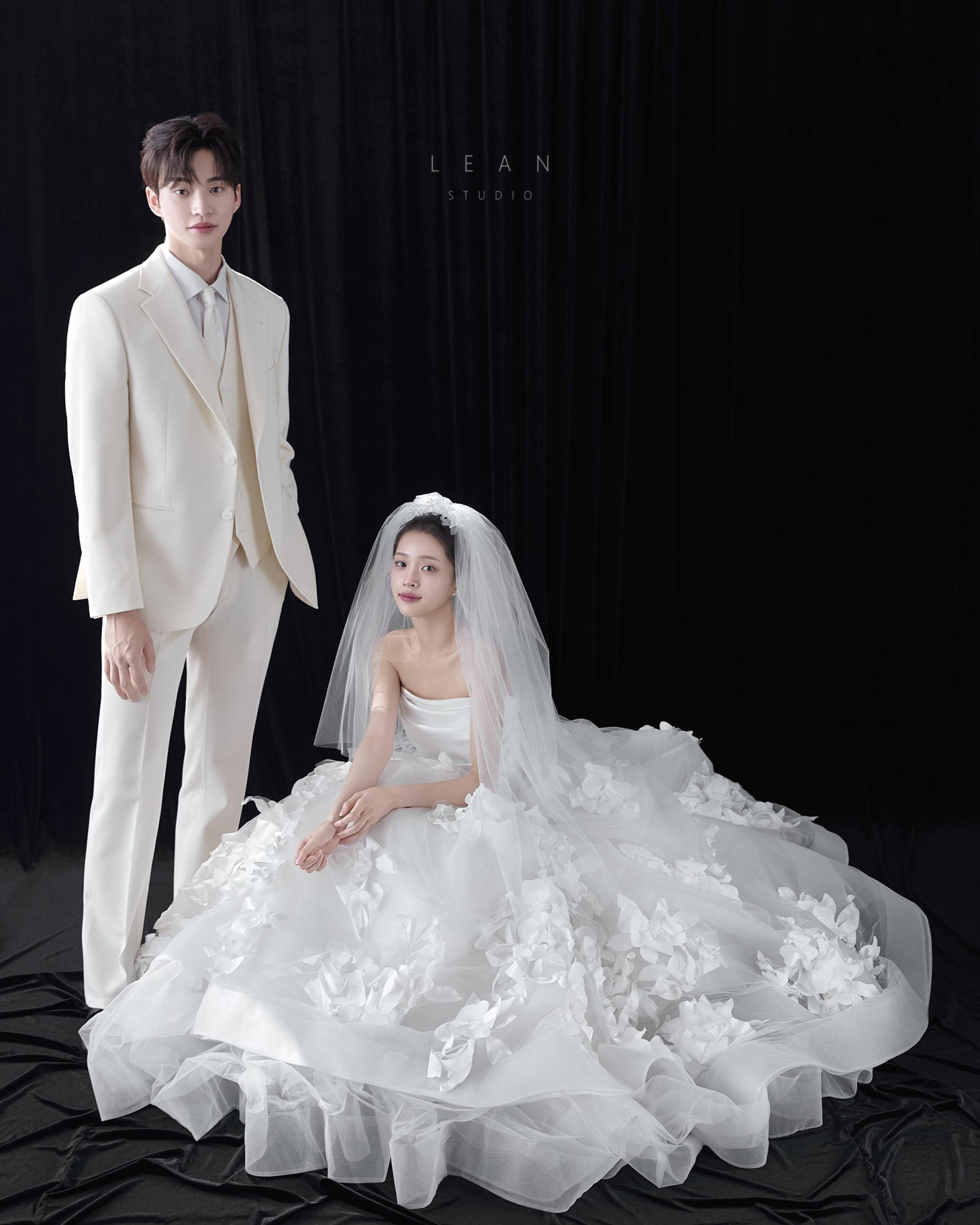 르안스튜디오 웨딩 스튜디오 블랙 커튼을 배경으로 화이트 수트 신랑과 입체 플라워 장식의 풍성한 웨딩드레스를 입고 베일을 쓴 신부를 대비감 있게 담은 웨딩 촬영컷