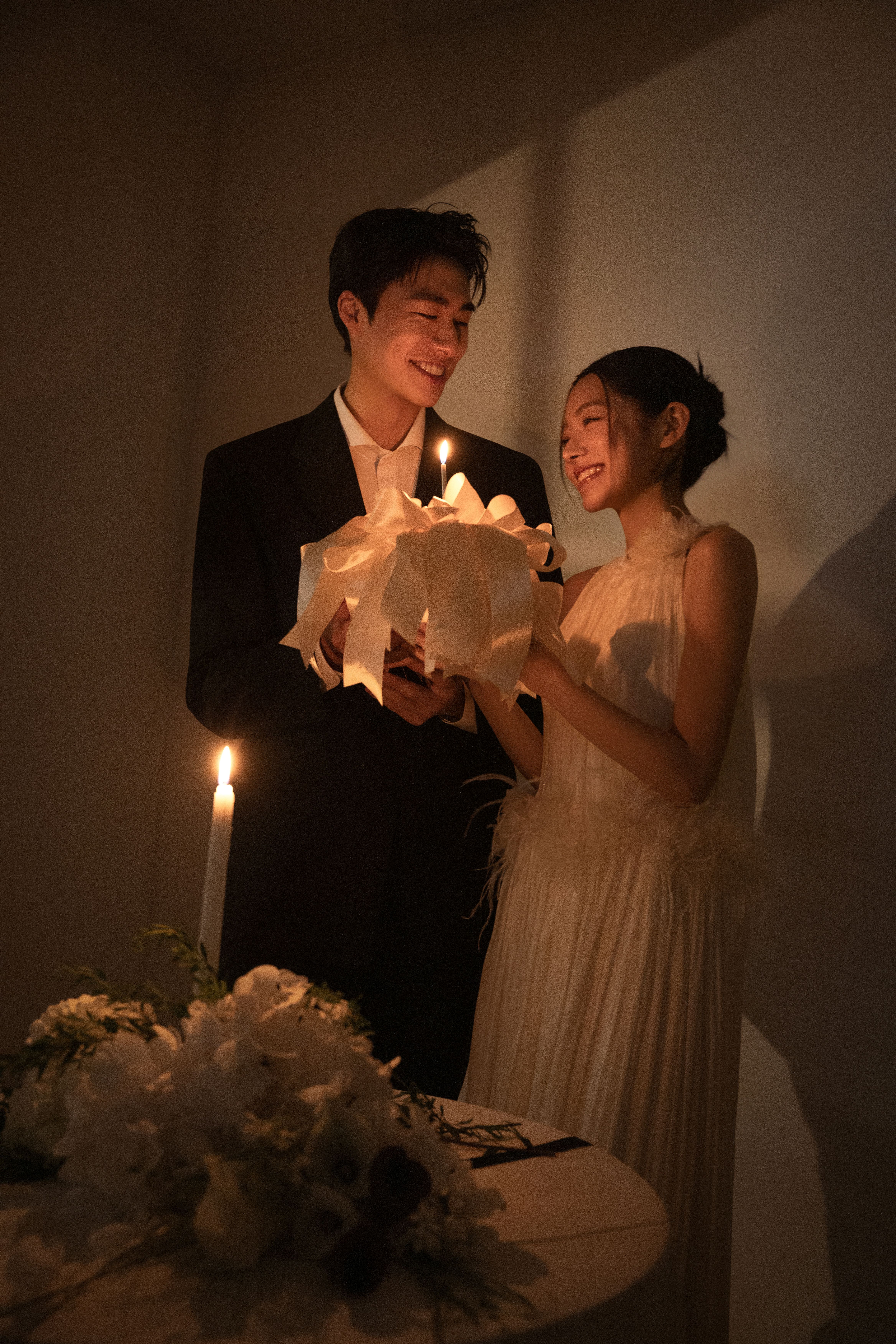 로렌하우스 웨딩 스튜디오 대표 컷 은은한 촛불 조명과 깊은 명암이 어우러진 실내에서 블랙 수트의 신랑과 홀터넥 웨딩드레스의 신부가 케이크를 함께 들고 서로를 바라보는 순간을 담아낸 따뜻하고 무드 있는 웨딩 촬영