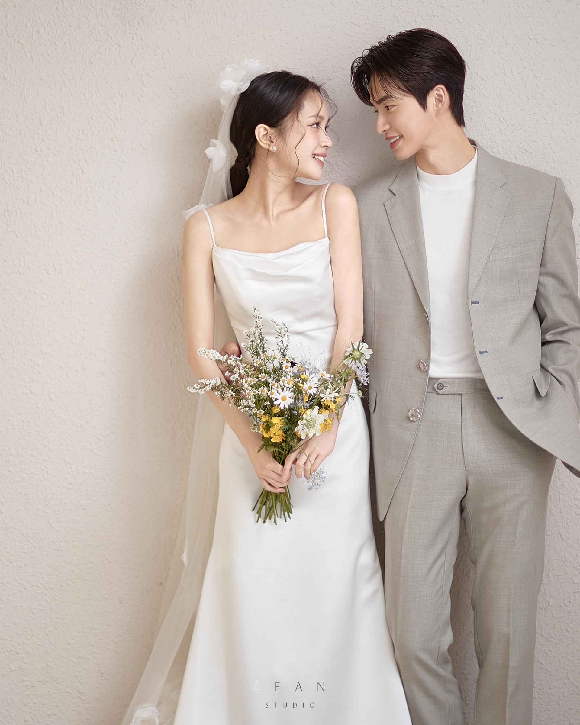 르안스튜디오 웨딩 스튜디오 화이트 벽면 앞에서 들꽃 부케를 든 슬립 웨딩드레스 신부와 라이트 그레이 수트 신랑이 서로 바라보며 미소 짓는 웨딩 촬영컷