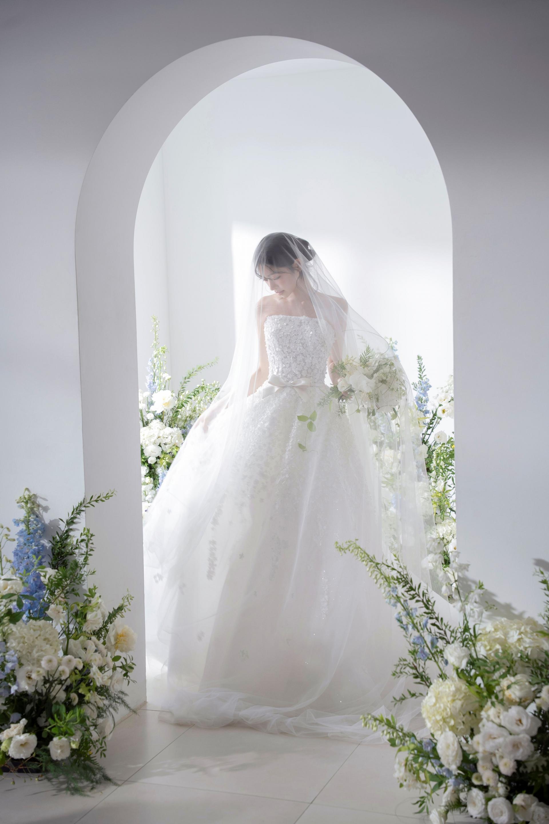 로렌하우스 웨딩 스튜디오 플라워 장식이 어우러진 아치형 공간에서 베일을 쓰고 화이트 부케를 든 신부 단독 웨딩 사진