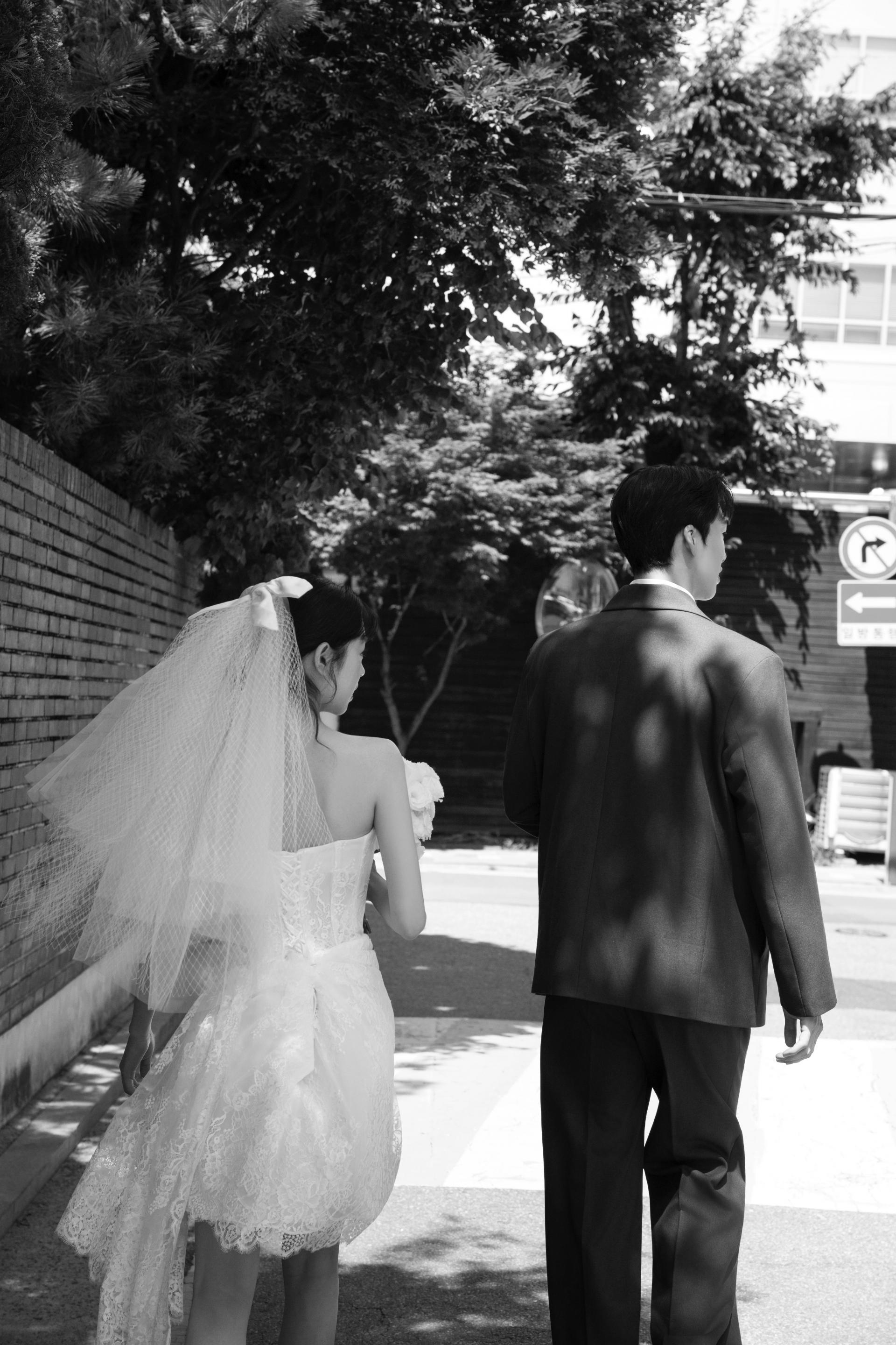 로렌하우스 웨딩 스튜디오 베일을 쓰고 부케를 든 신부와 수트를 입은 신랑이 야외 길을 나란히 걷는 모습을 흑백 감성으로 담은 커플 웨딩 사진