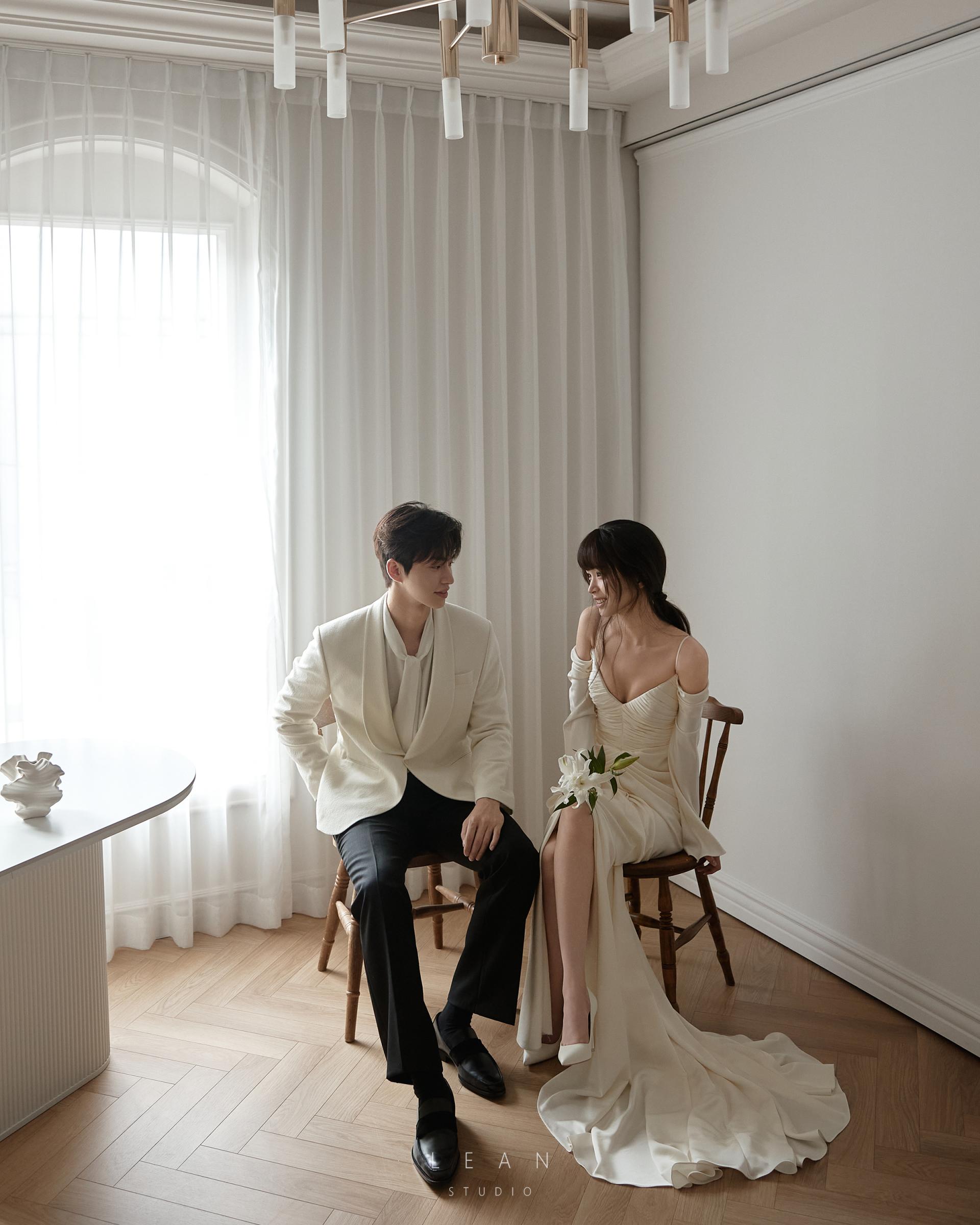 르안 스튜디오 웨딩 스튜디오 아치형 창가의 자연광이 스며드는 화이트 인테리어에서 우드 체어에 나란히 앉아 서로를 바라보는 신랑 신부의 웨딩 촬영컷