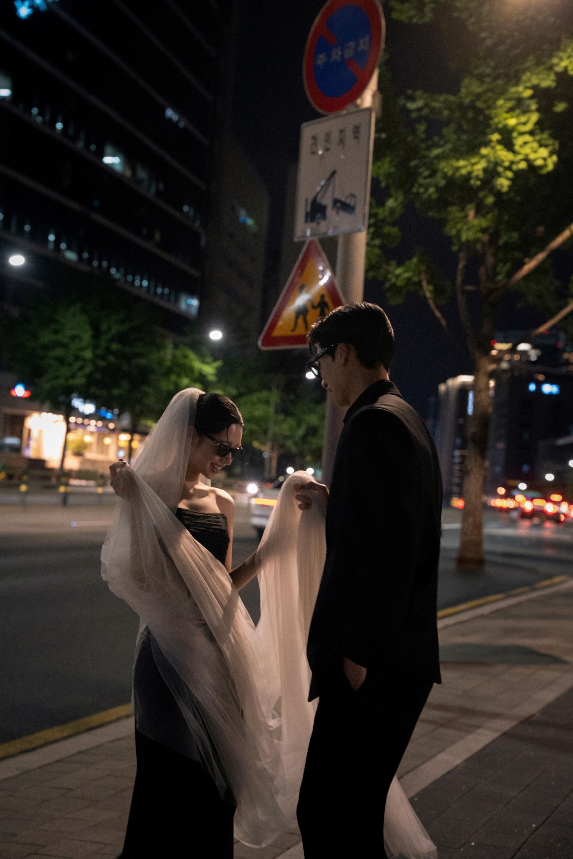 로렌하우스 웨딩 스튜디오 야외 밤거리에서 선글라스를 끼고 블랙 드레스와 수트를 입은 신랑 신부가 베일을 만지며 미소 짓는 커플 웨딩 사진