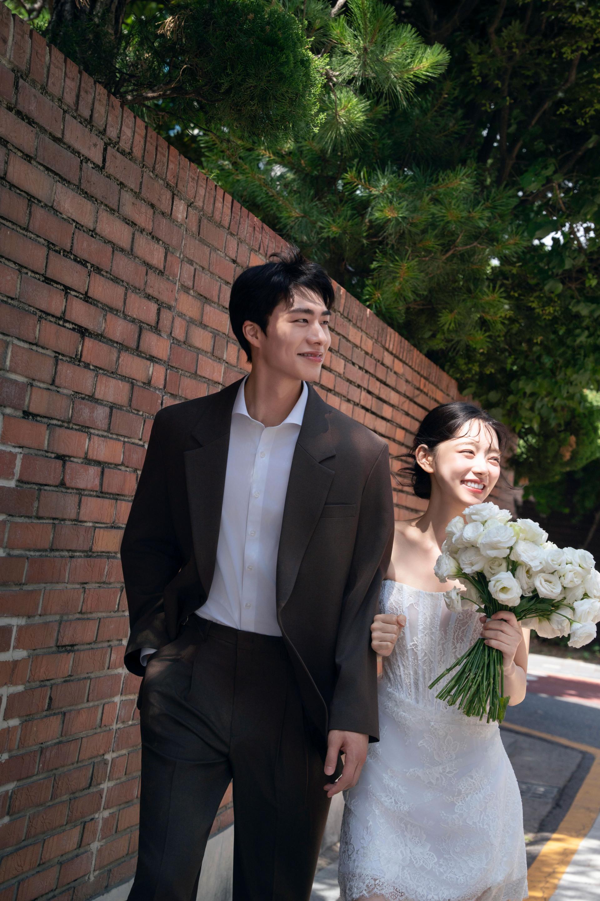 로렌하우스 웨딩 스튜디오 붉은 벽돌 담장을 배경으로 브라운 수트를 입은 신랑과 화이트 부케를 들고 걷는 신부의 야외 웨딩 사진