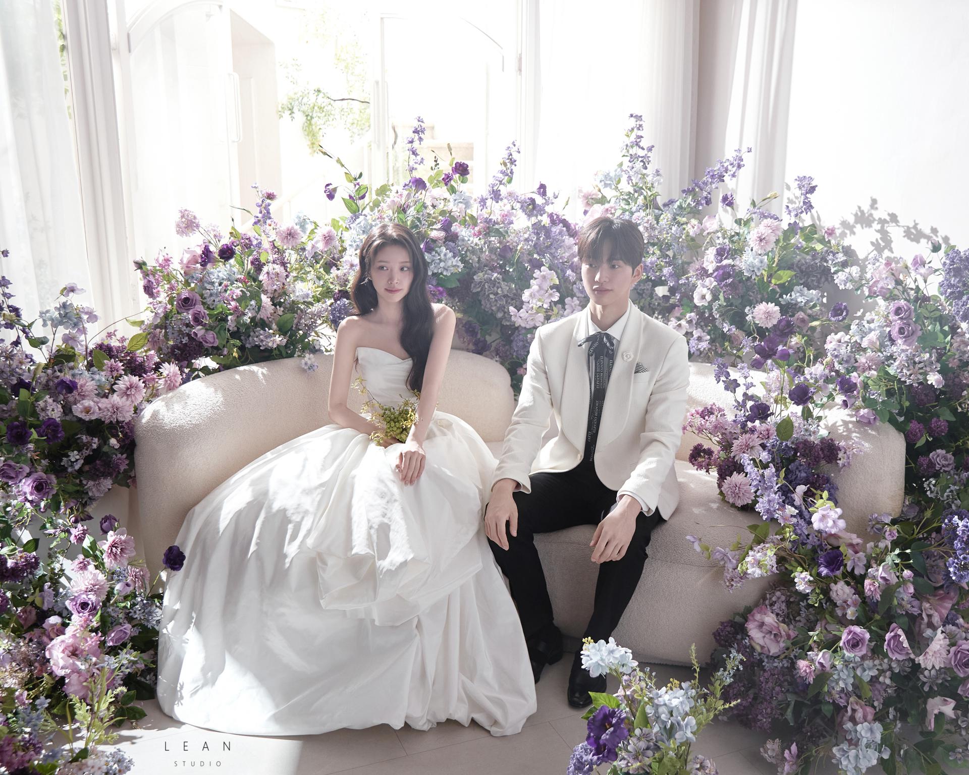 르안스튜디오 웨딩 스튜디오 자연광이 쏟아지는 창가에서 퍼플과 라벤더 컬러 플라워로 둘러싸인 소파에 웨딩드레스 신부와 화이트 재킷 신랑이 나란히 앉은 플라워씬 웨딩 촬영컷