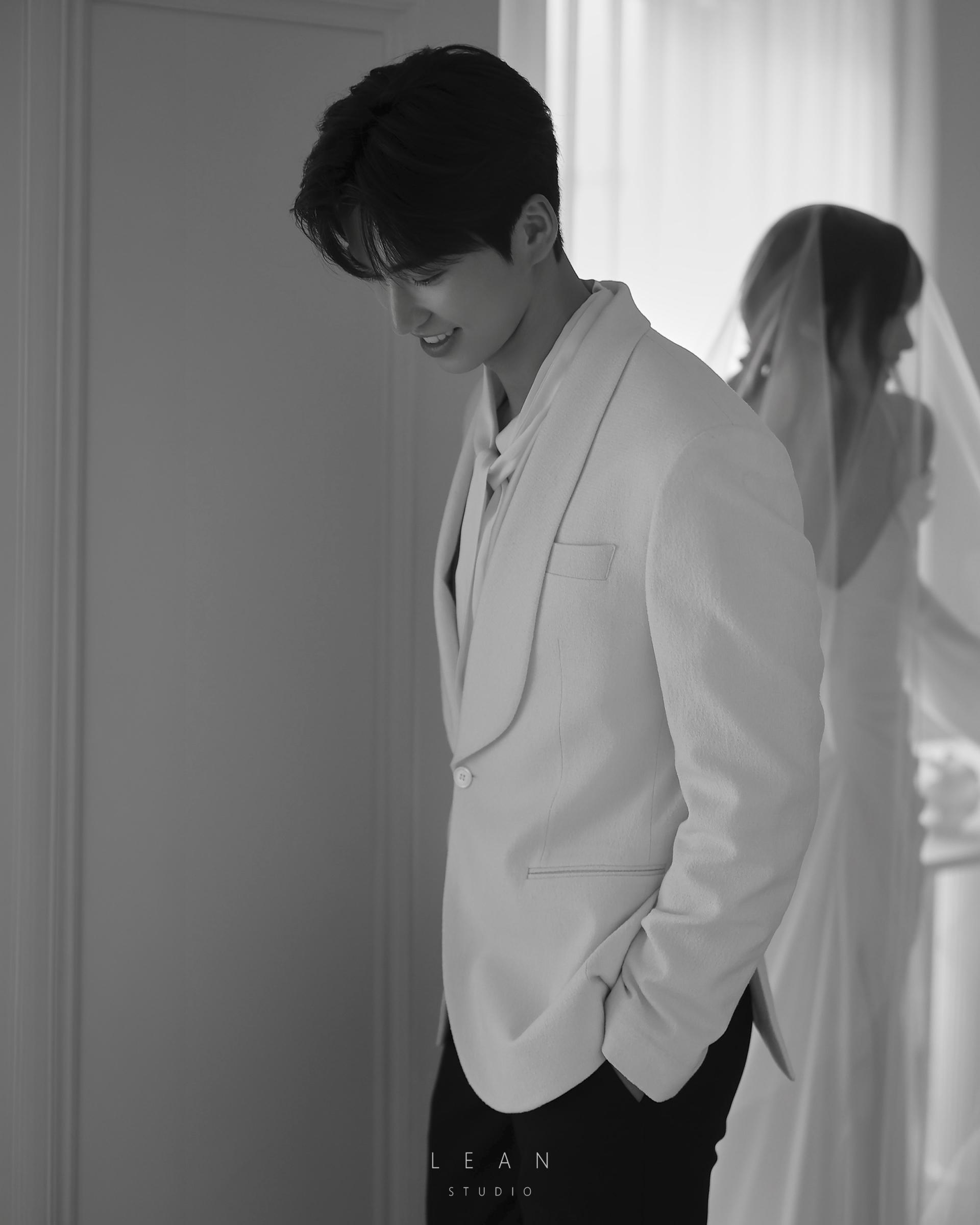 르안스튜디오 웨딩 스튜디오 밝은 창가를 배경으로 화이트 재킷을 입은 신랑과 롱 베일을 쓴 신부를 앞뒤 구도로 담은 감성적인 흑백 웨딩 촬영컷