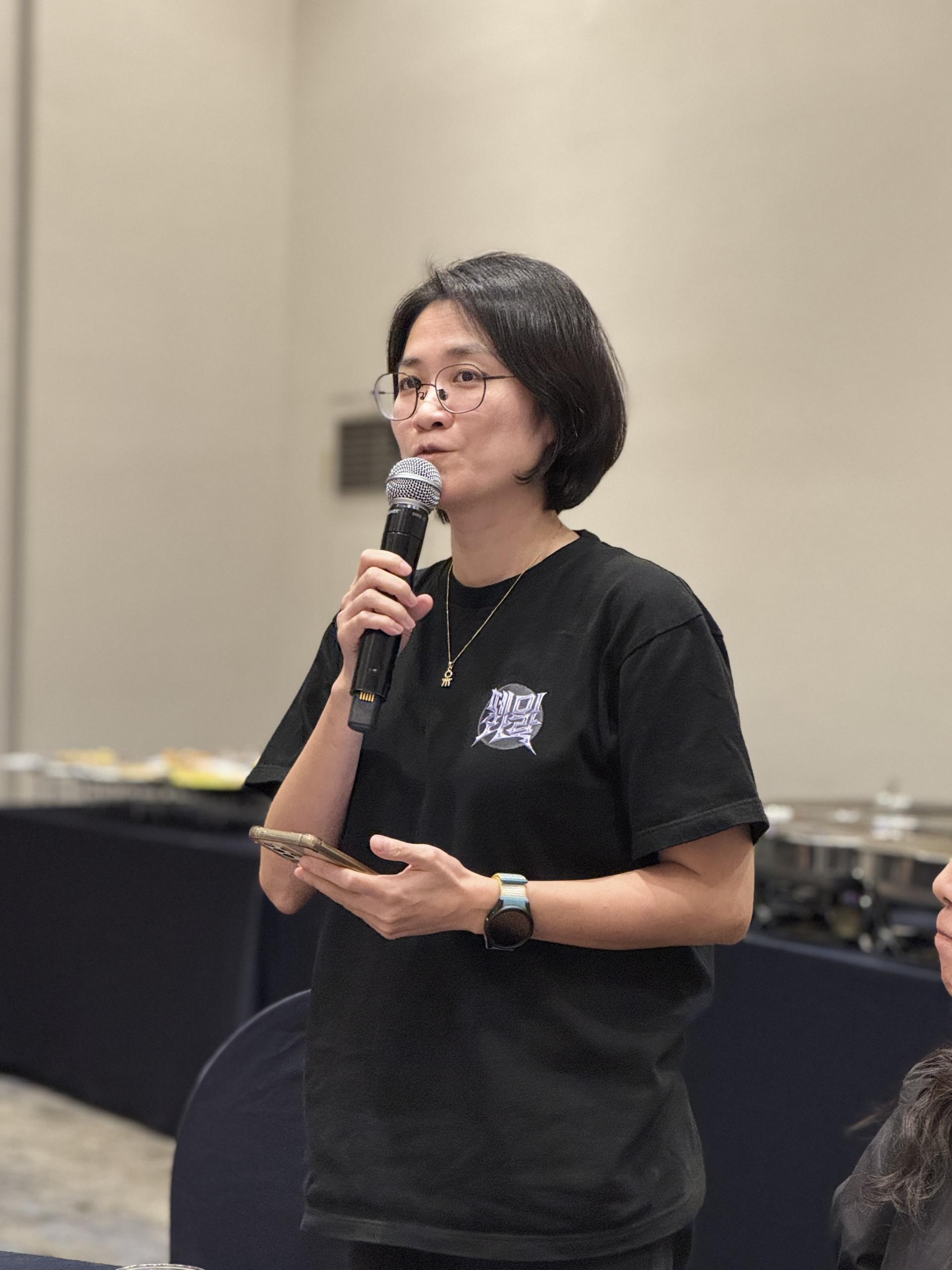 검은 옷을 입고 안경을 쓴 한 사람이 일어나 핸드폰과 마이크를 양 손에 들고 진지한 표정으로 뭔가 말하고 있다. 뒤쪽엔 뷔페 식사 라인이 보인다. 