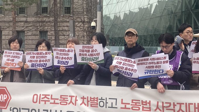 기자회견 참가자들이 심각한 얼굴로 피켓을 들고 서있다. 가운데 안경을 쓴 단발머리 여성이 발언하고 있다.