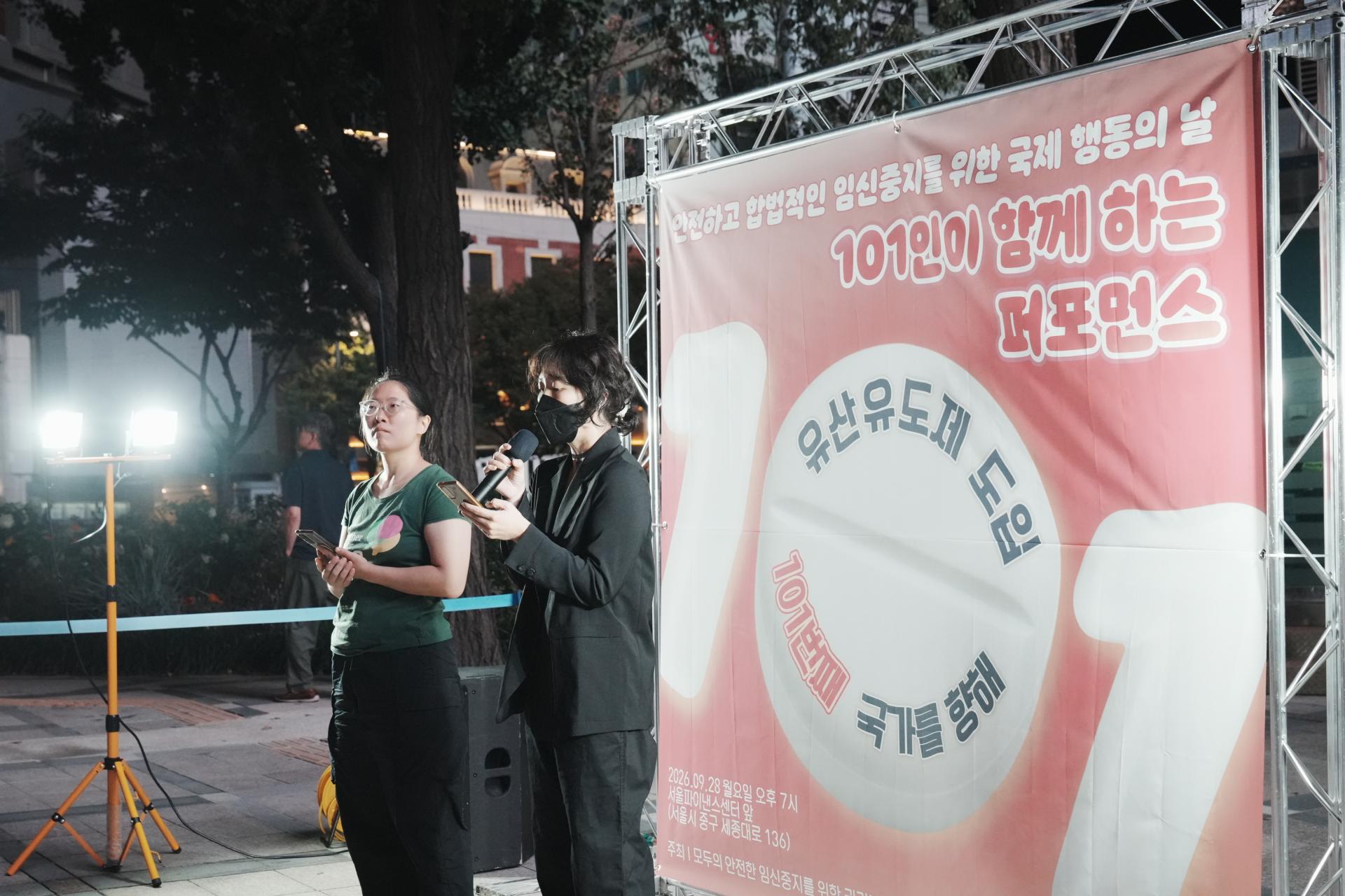 검은 마스크를 끼고, 검은색 상하의를 맞춰 입은 시민이 핸드폰을 바라보며 발언하고 있다. 그 옆으로는 또 다른 시민참여자가 핸드폰을 든 채 순서를 기다리고 있다.