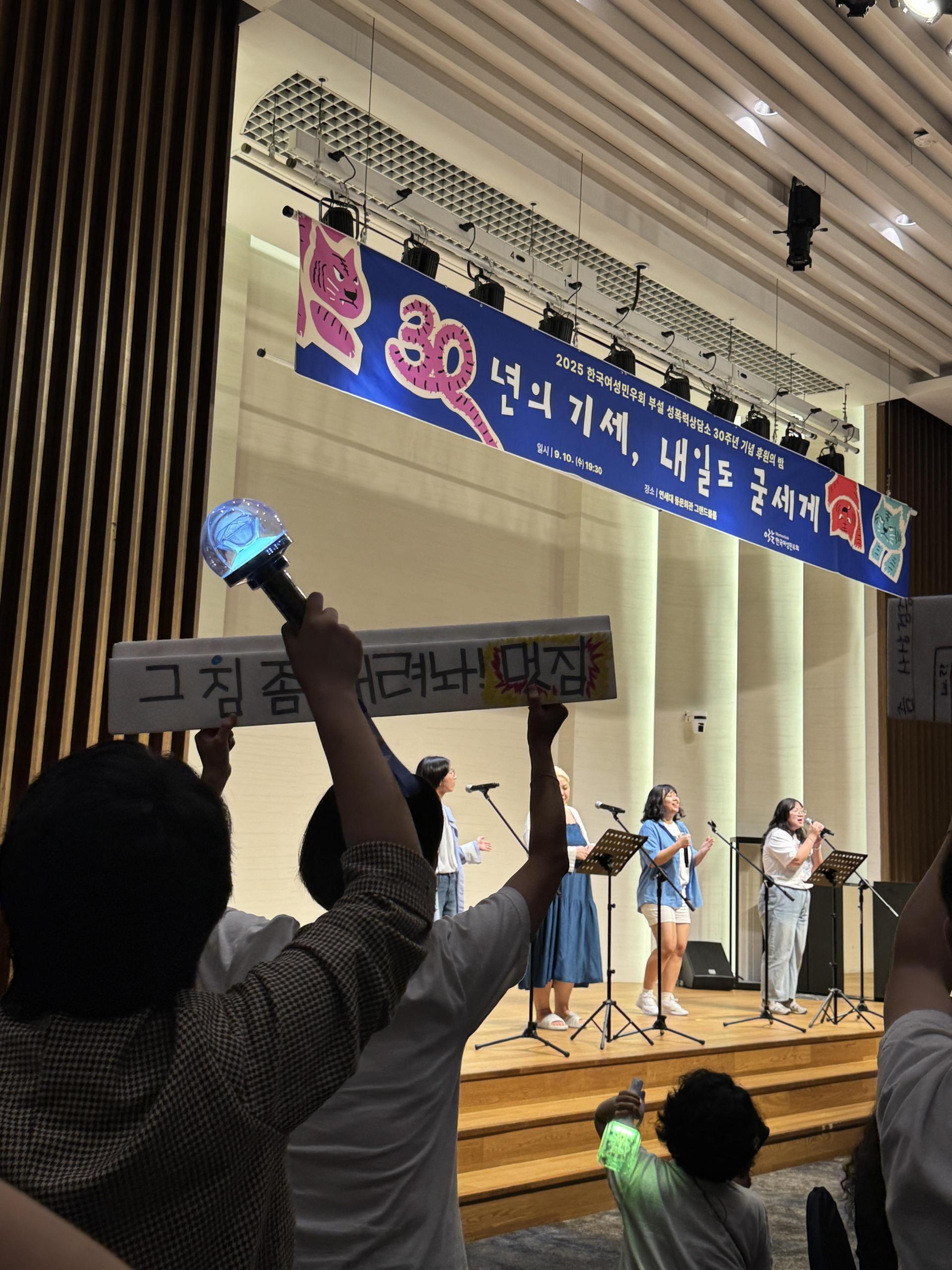 무대엔 네 명의 사람이 공연하고 있고, 무대 아래에서 무대를 향해 응원봉과 응원피켓을 흔들고 있는 여러 사람의 뒷모습이 보인다. 무대 위 현수막이 가운데쯤 보이는데 파란 배경에 30년의 기세, 내일도 굳세게 라고 적혀 있다.