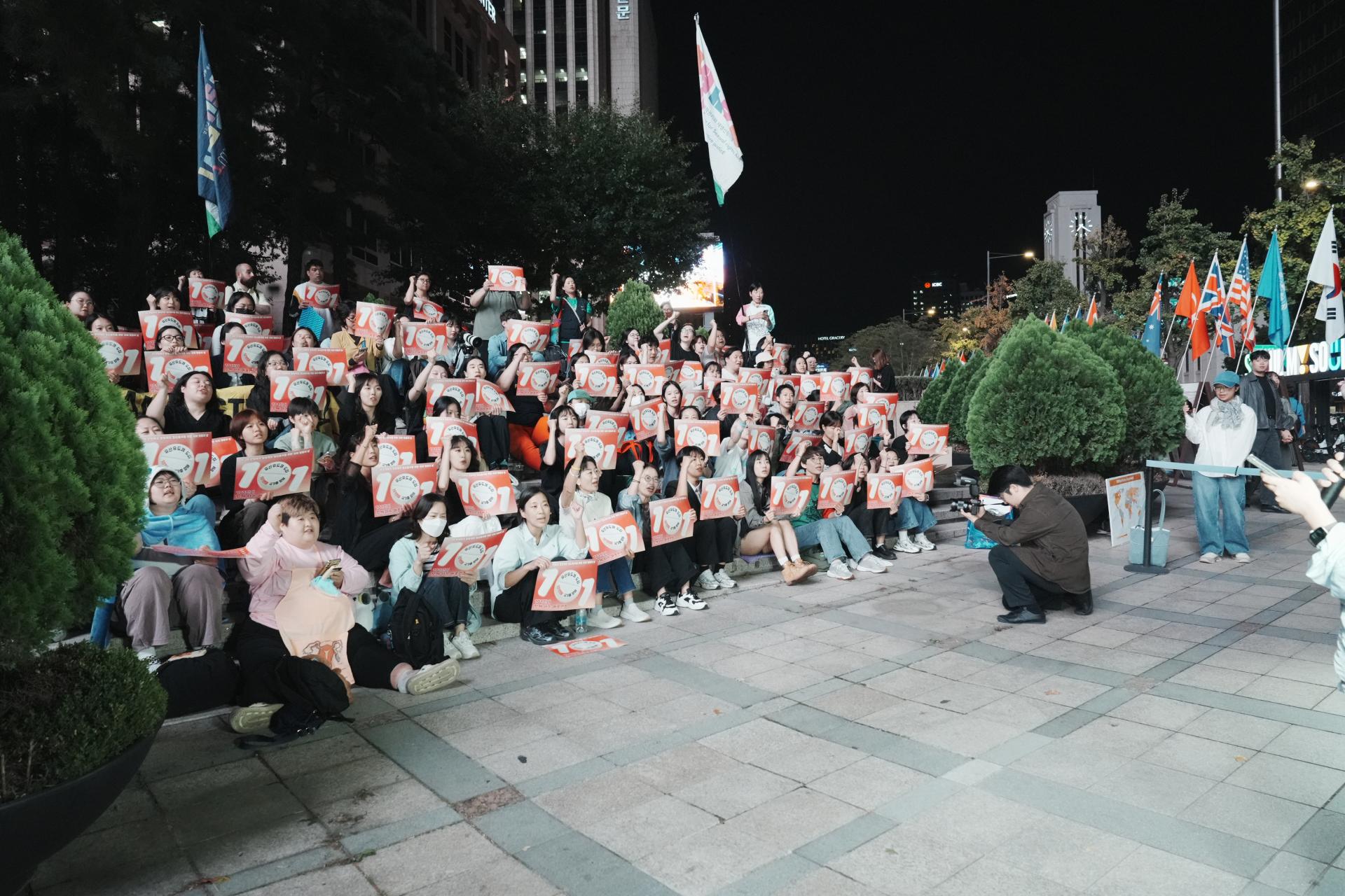 한 손으로는 손피켓을, 다른 한 손으로는 오른손을 위로 번쩍 든 참여자들의 모습. 참여자들은 화분 뒤쪽으로 위치한 계단에 앉아있고, 모두 밝은 얼굴을 한 채 구호를 외치고 있다.