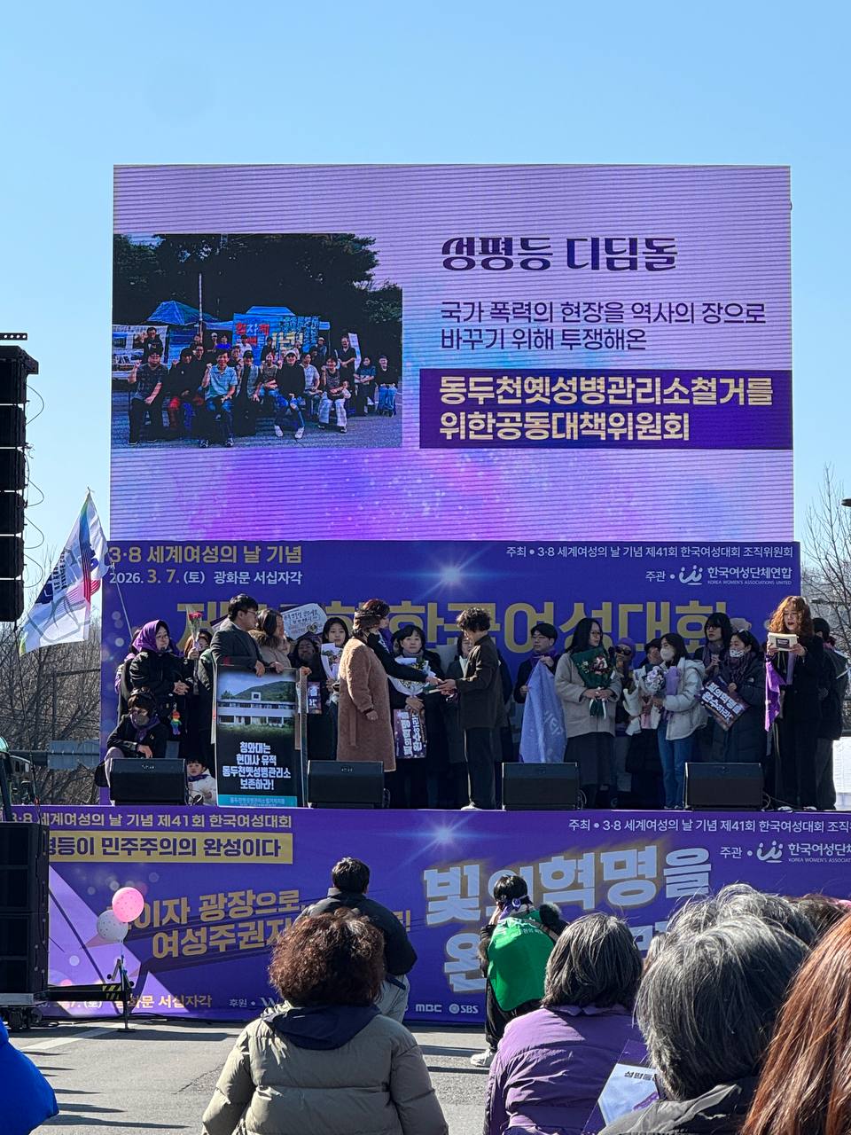 사진 33. 무대에서 올해의 성평등 디딤돌을 발표하고 있다. 수상자 이름 영화 동두천옛성병관리소철거저지를 위한 공동대책위원회가 무대 스크린에 게시되어있다. 사진에는 동두천옛성병관리소철거를위한공동대책위원회로 쓰여있으나, 철거가 아닌 철거저지를 위한 공동대책위원회이다.