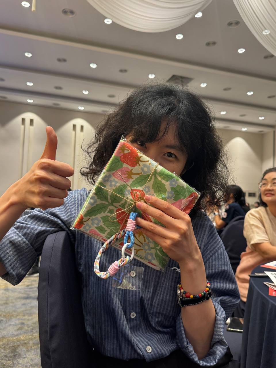 줄무늬 셔츠를 입은 한 사람이 알록달록하게 포장된 선물로 얼굴 아랫쪽을 반쯤 가리고, 카메라를 향해 엄지를 들어올리는 모습. 눈이 웃고 있다. 