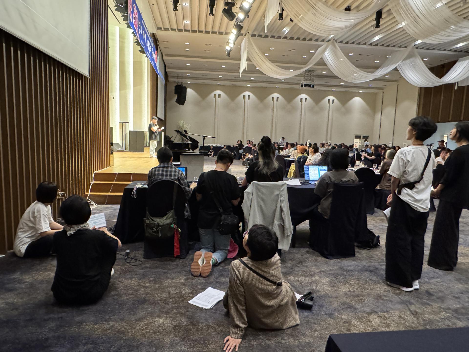 "넓은 행사장. 무대 옆 행사진행 스탭 테이블 뒤쪽의 모습. 사람들이 앉거나 서서 무대 위 발표자를 바라보고 있다. 몇몇은 바닥에 앉아 있고, 이들 너머에는 원형 테이블에 앉은 관객들이 무대를 향해 보고 있다. 천장에는 흰 천 장식이 걸려 있다."